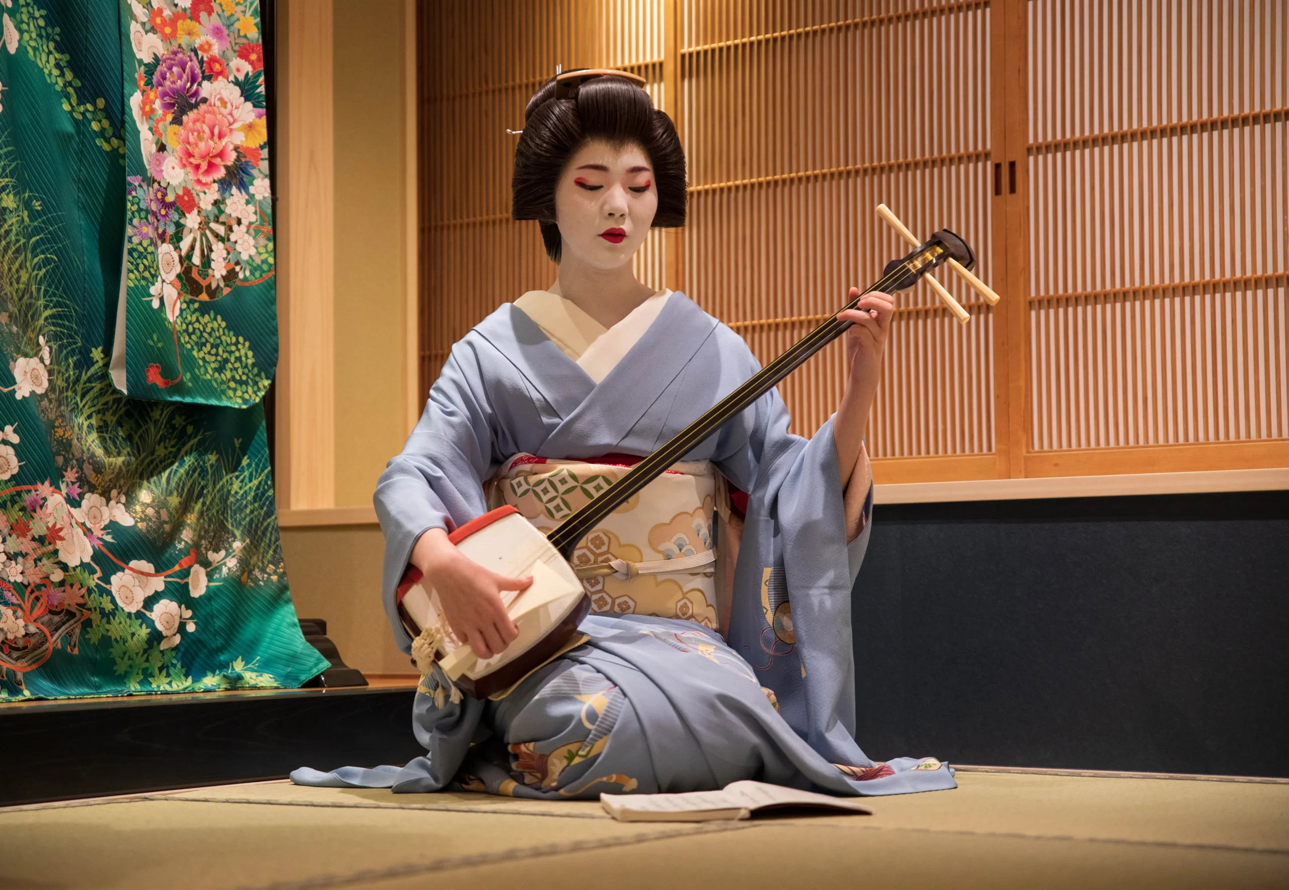 A geisha dressed in a light blue kimono gracefully plays the shamisen in a traditional Japanese tatami room with wooden interiors and a decorative fabric backdrop.