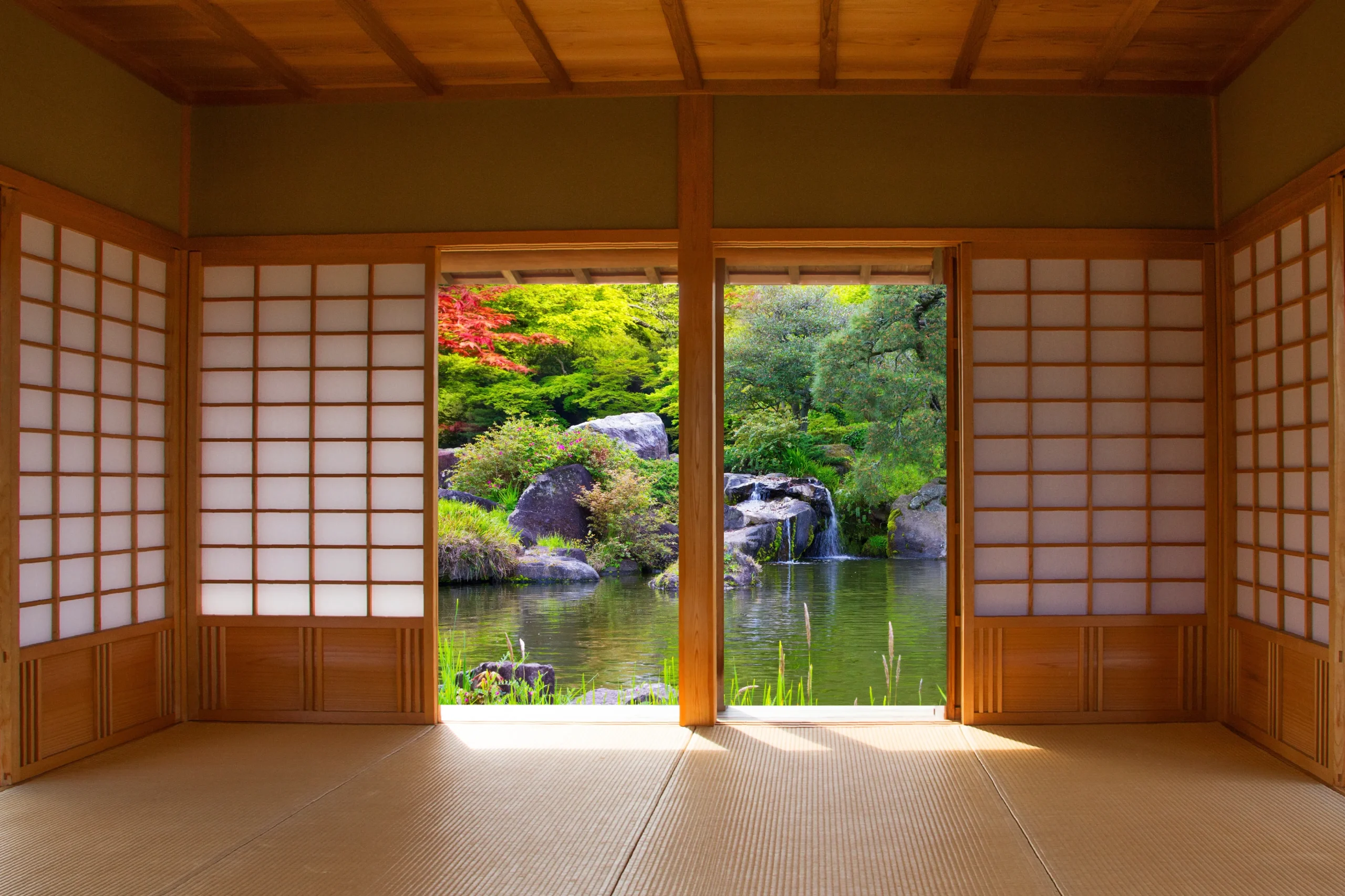 A serene view from inside a traditional Japanese tatami room, looking out through sliding shoji doors onto a beautiful Japanese garden with lush greenery, a pond, and a small waterfall.