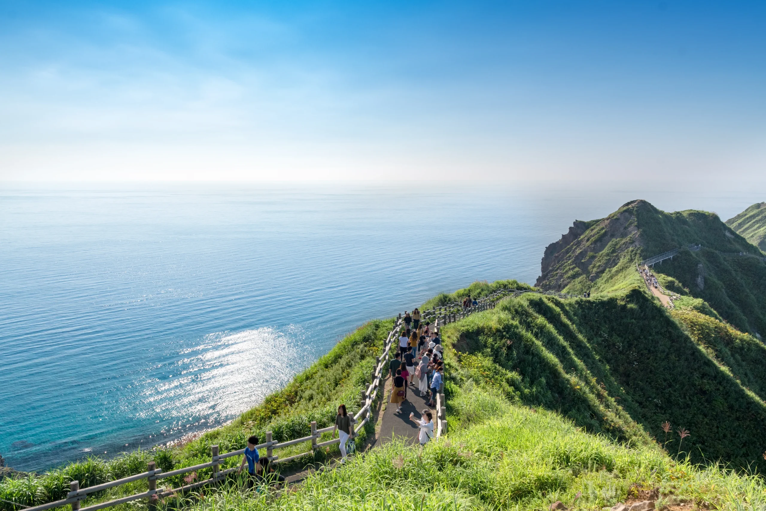 Visitors walking along a narrow, scenic trail at Cape Kamui in Hokkaido, with sweeping views of the turquoise sea and rugged cliffs under a clear blue sky.