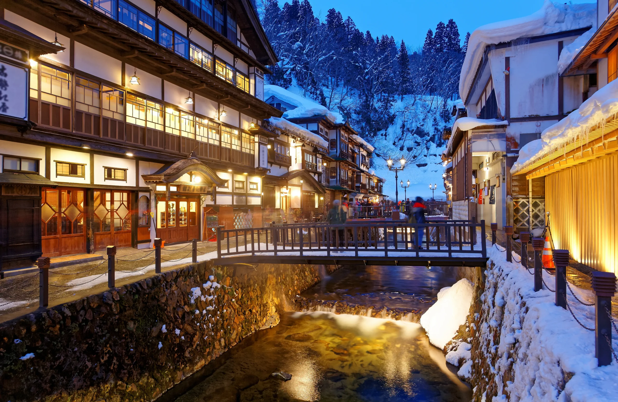 A picturesque hot spring town during winter, featuring snow-covered wooden buildings and a bridge over a stream, creating a magical evening atmosphere