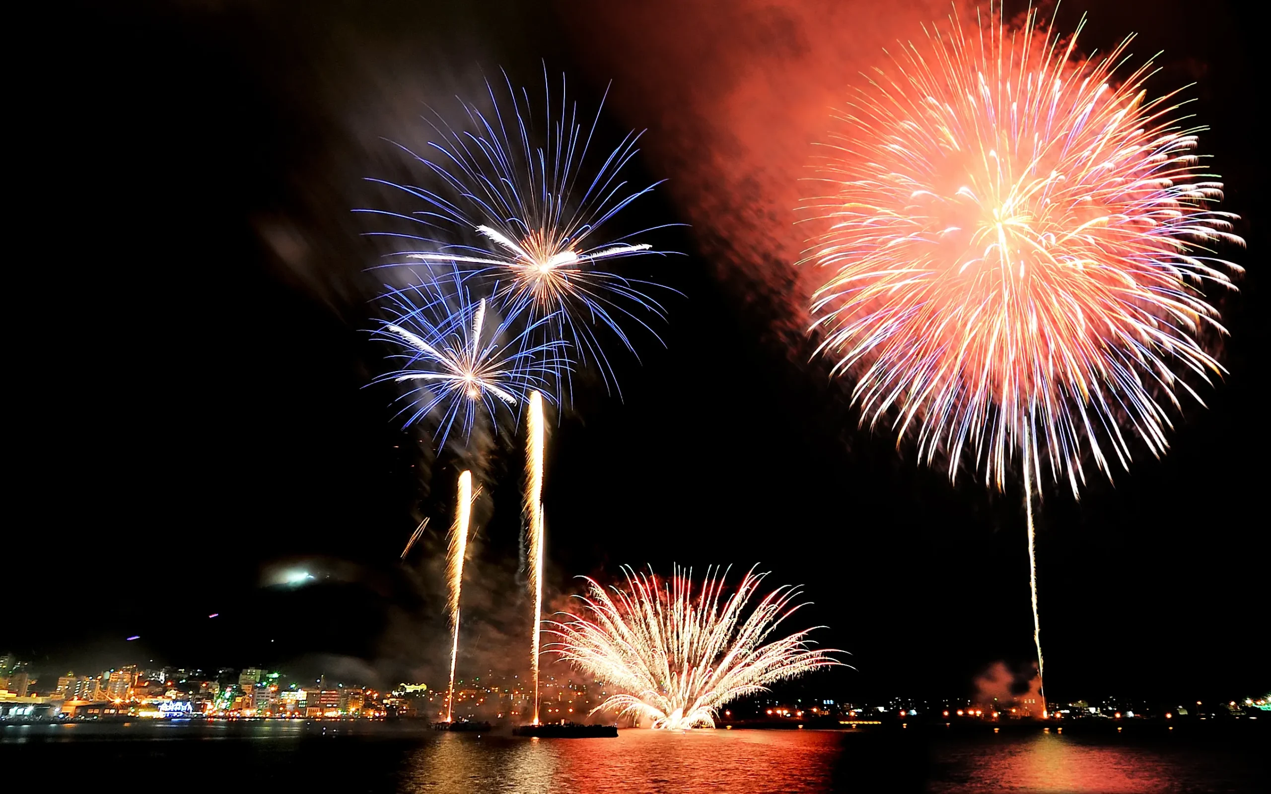 A stunning display of fireworks in vibrant red and blue colors illuminating the night sky over Hakodate Port, with the city lights reflecting on the water