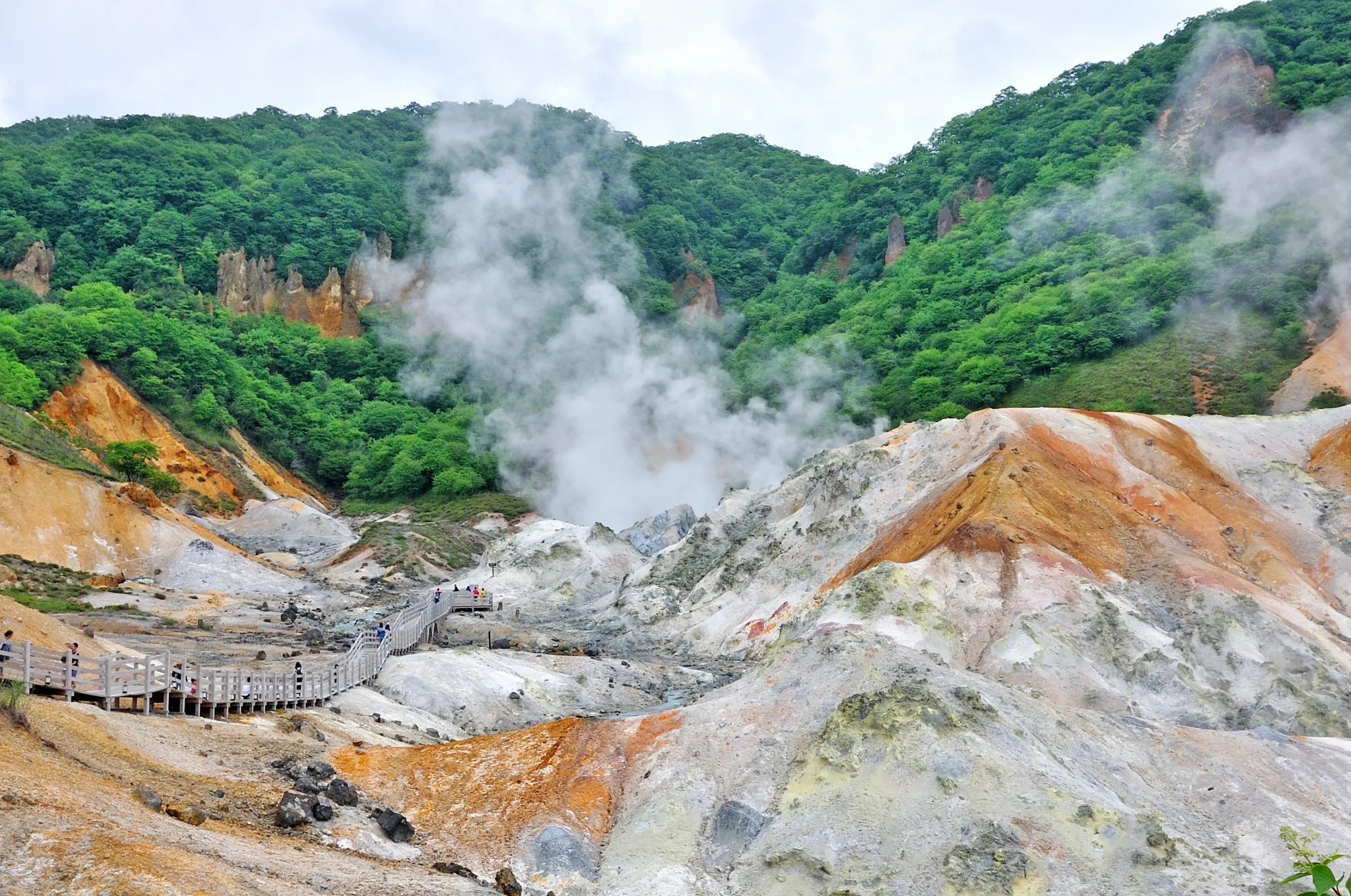 Steaming volcanic valley with sulfur vents, rocky terrain, and a wooden walkway surrounded by lush green forested hills.