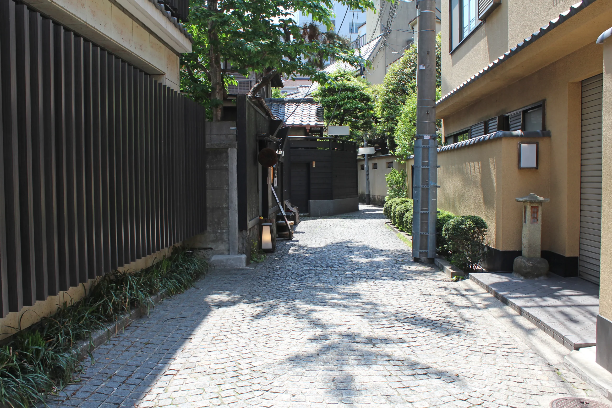 A traditional Japanese neighborhood with a stone-paved street surrounded by wooden fences and well-maintained greenery, offering a serene and nostalgic feel.