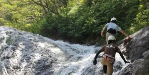 Two people climbing a cascading hot spring waterfall surrounded by lush green forest, wearing helmets for safety, at Kamuiwakka Hot Falls in Shiretoko, Hokkaido.