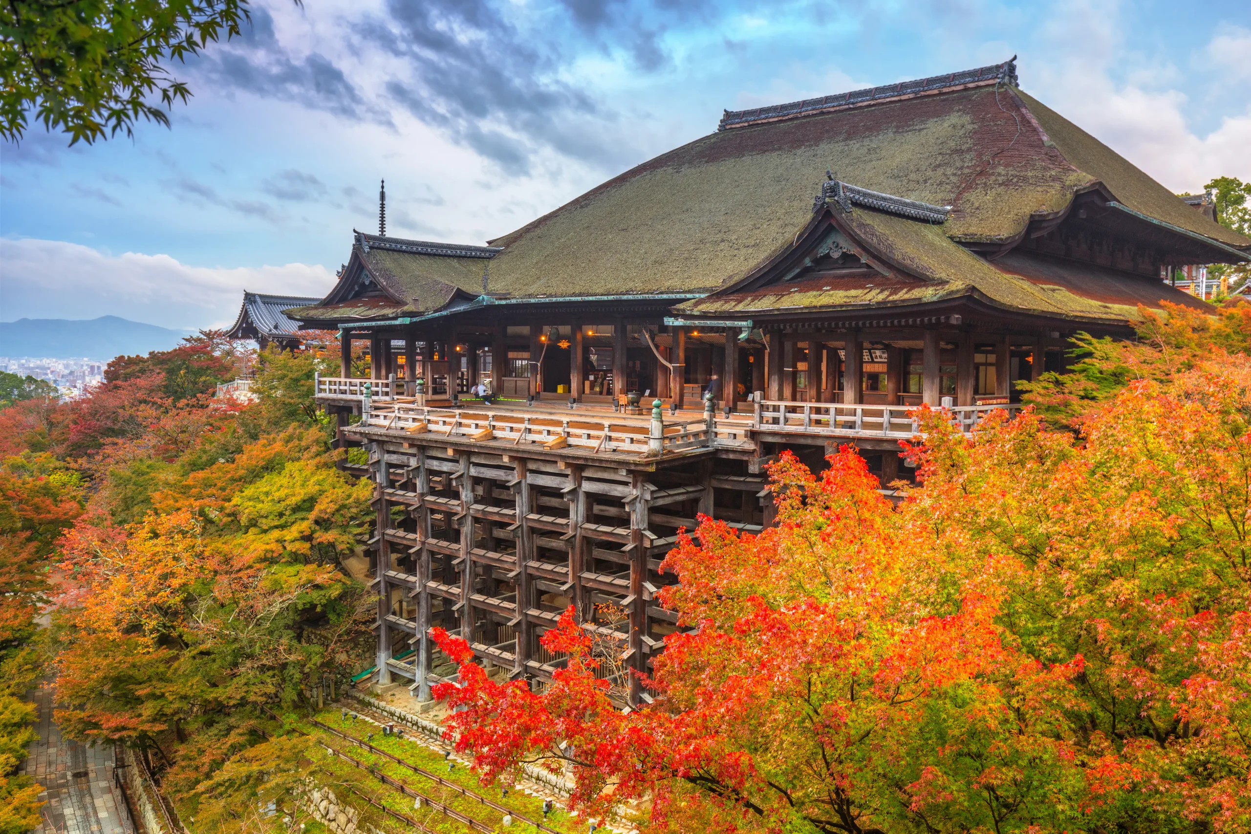 Kiyomizu-dera Temple in Kyoto, perched on wooden stilts above a hillside of fiery autumn foliage, under a partly cloudy sky.
