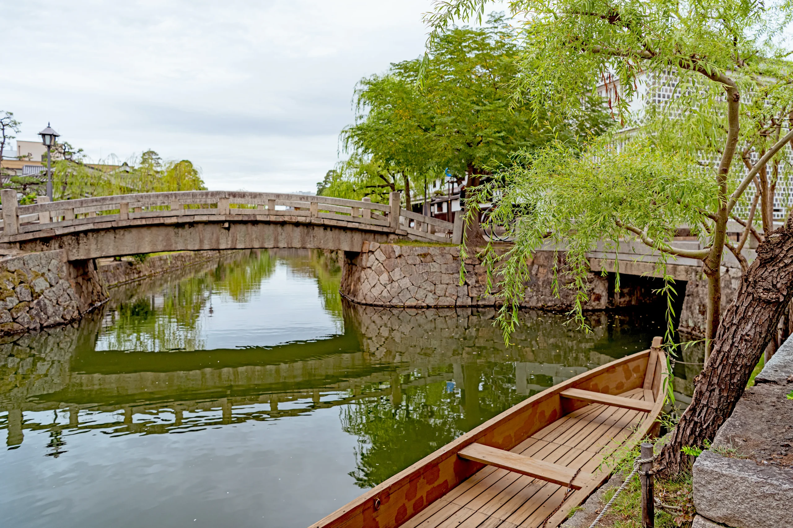 A tranquil canal lined with willow trees and traditional Japanese architecture, including a small wooden boat and a stone bridge.