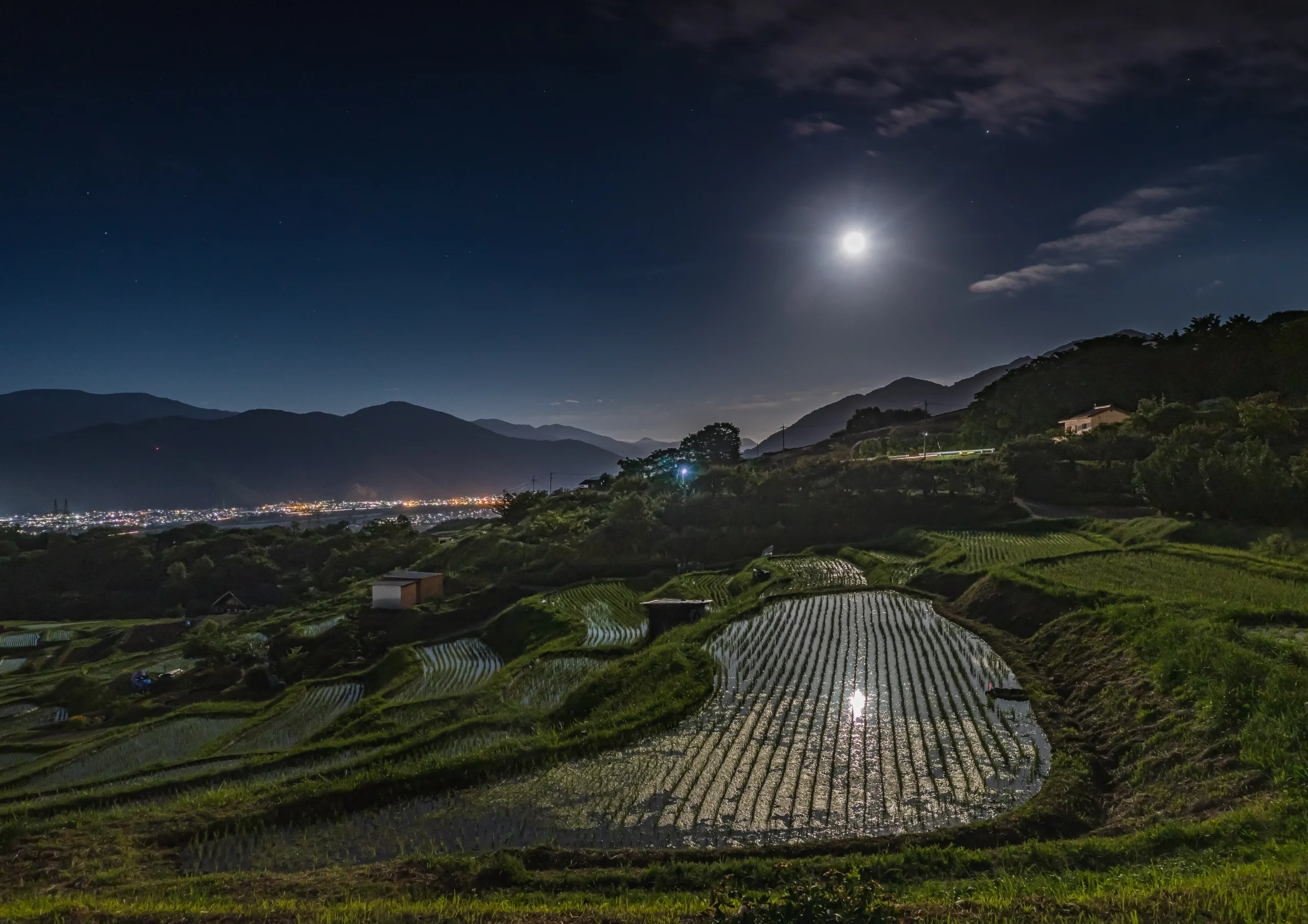 A tranquil night view of the Obasute rice fields, with a glowing full moon casting light on the reflective water, surrounded by distant city lights and mountains.