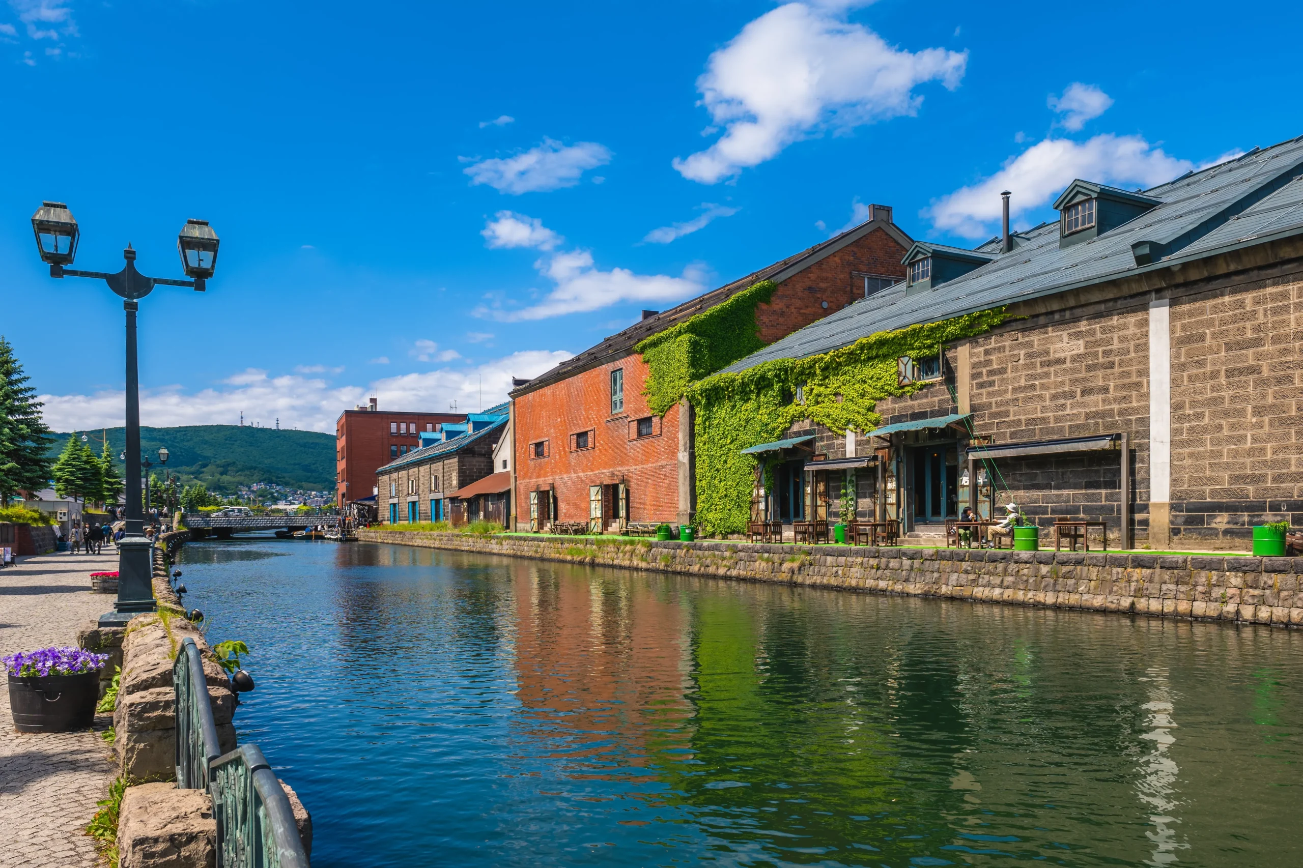 A scenic canal lined with red-brick warehouses, some covered in ivy, under a bright blue sky, reflecting the historic charm of Otaru.