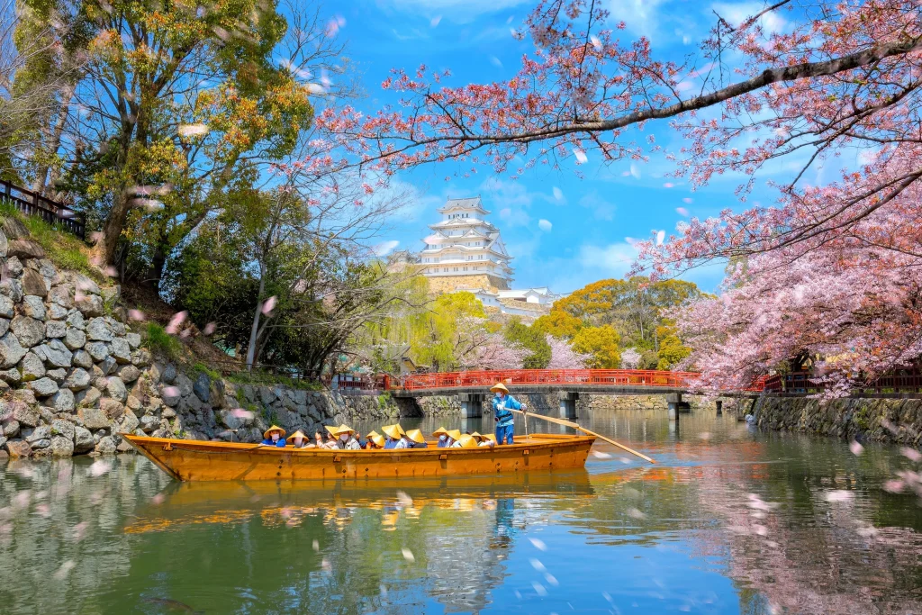 A boat glides along a canal lined with cherry blossoms, with Himeji Castle standing in the background.