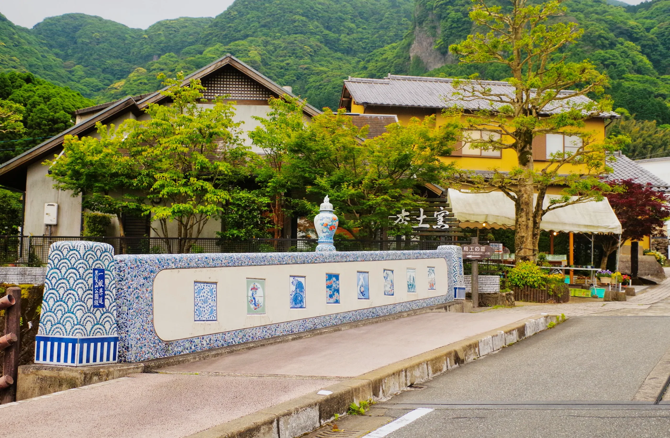 A picturesque street in Imari, Saga Prefecture, featuring a decorative bridge adorned with traditional porcelain designs, surrounded by lush greenery and traditional Japanese houses.