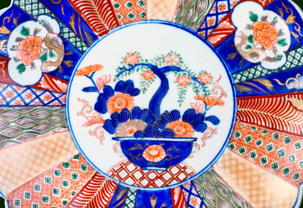 A detailed view of an Imari ware plate featuring a central floral motif with a tree in vivid blue and orange, surrounded by intricate geometric and floral patterns in bold colors.