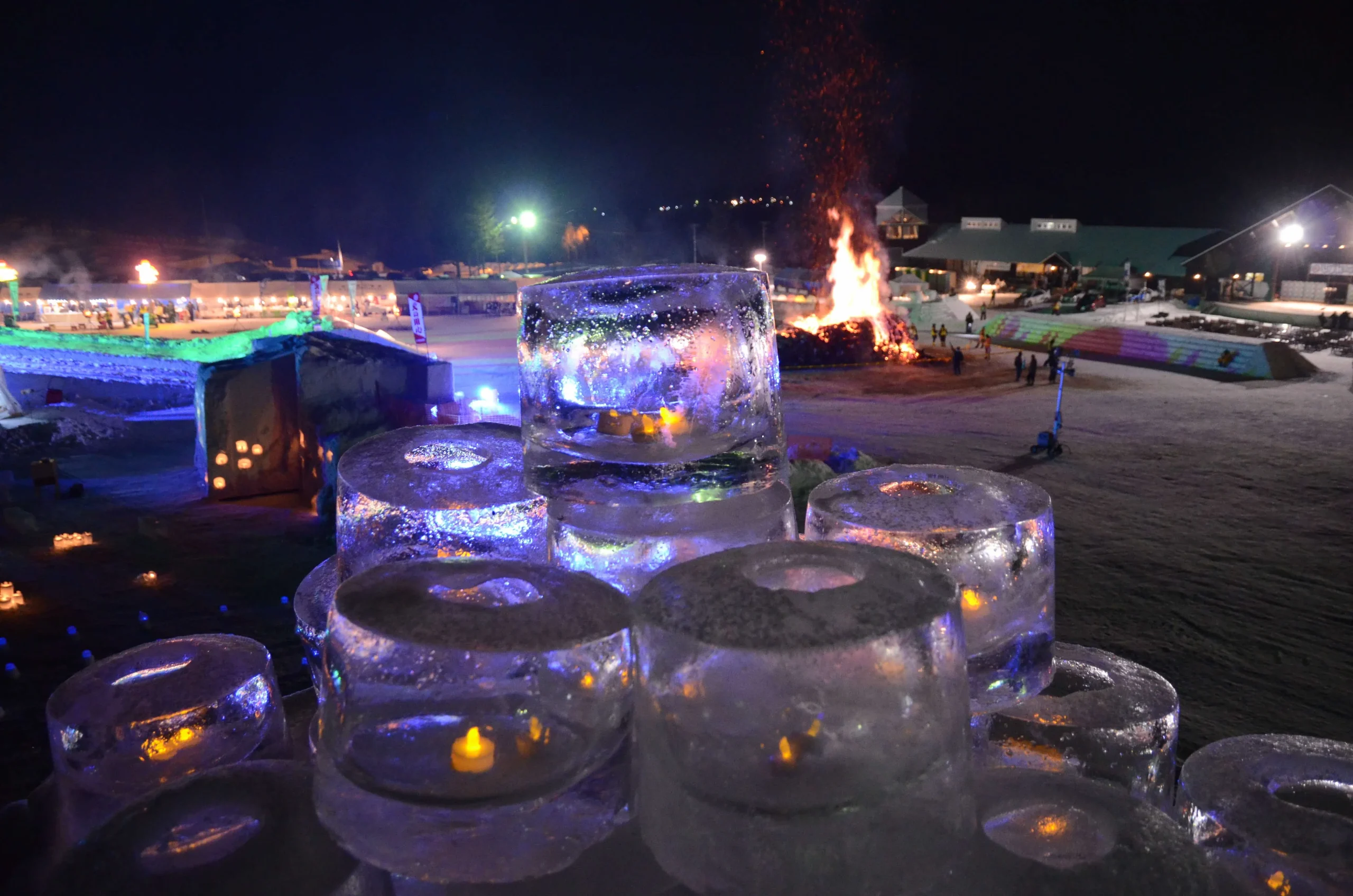 A night scene from the Rikubetsu Shibare Festival featuring glowing ice lanterns, a large bonfire, and illuminated winter decorations.