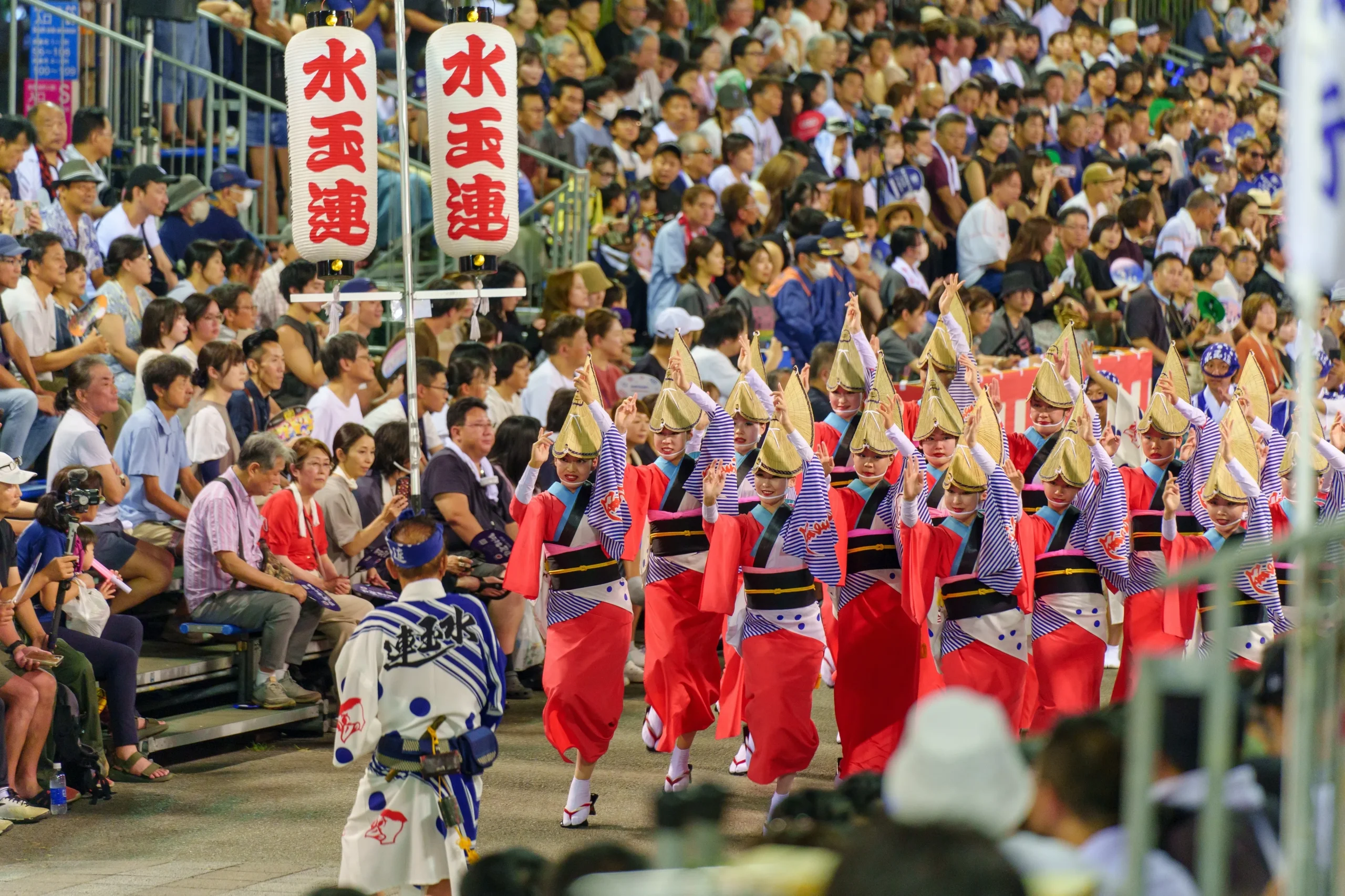 A lively Awa Odori dance performance in Tokushima, featuring female dancers in red and white kimono with straw hats, raising their hands in synchronized movements as they parade through a crowded street.