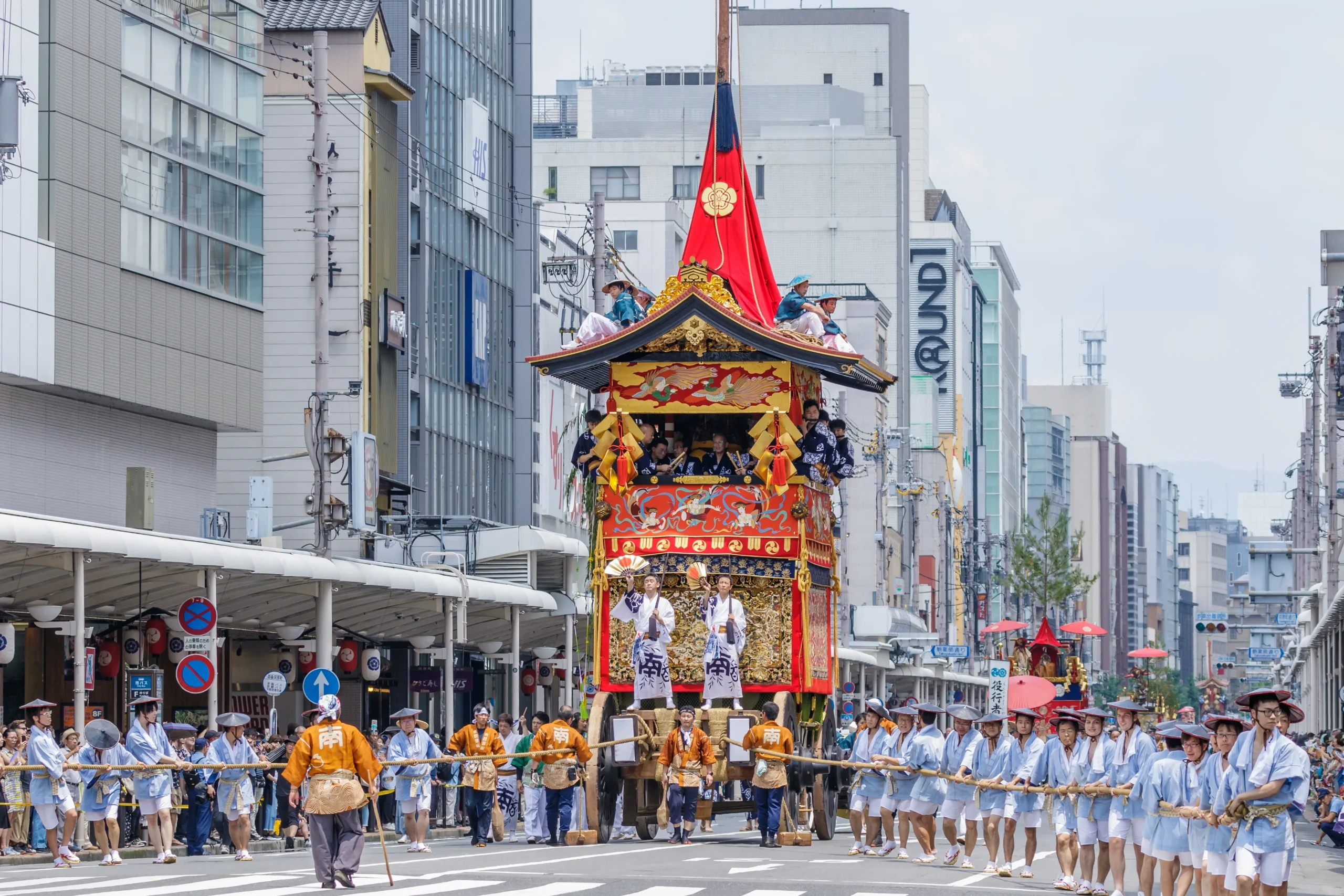 A grand float from the Gion Festival in Kyoto, adorned with intricate decorations and a red canopy, being pulled by participants in traditional attire through the city streets.