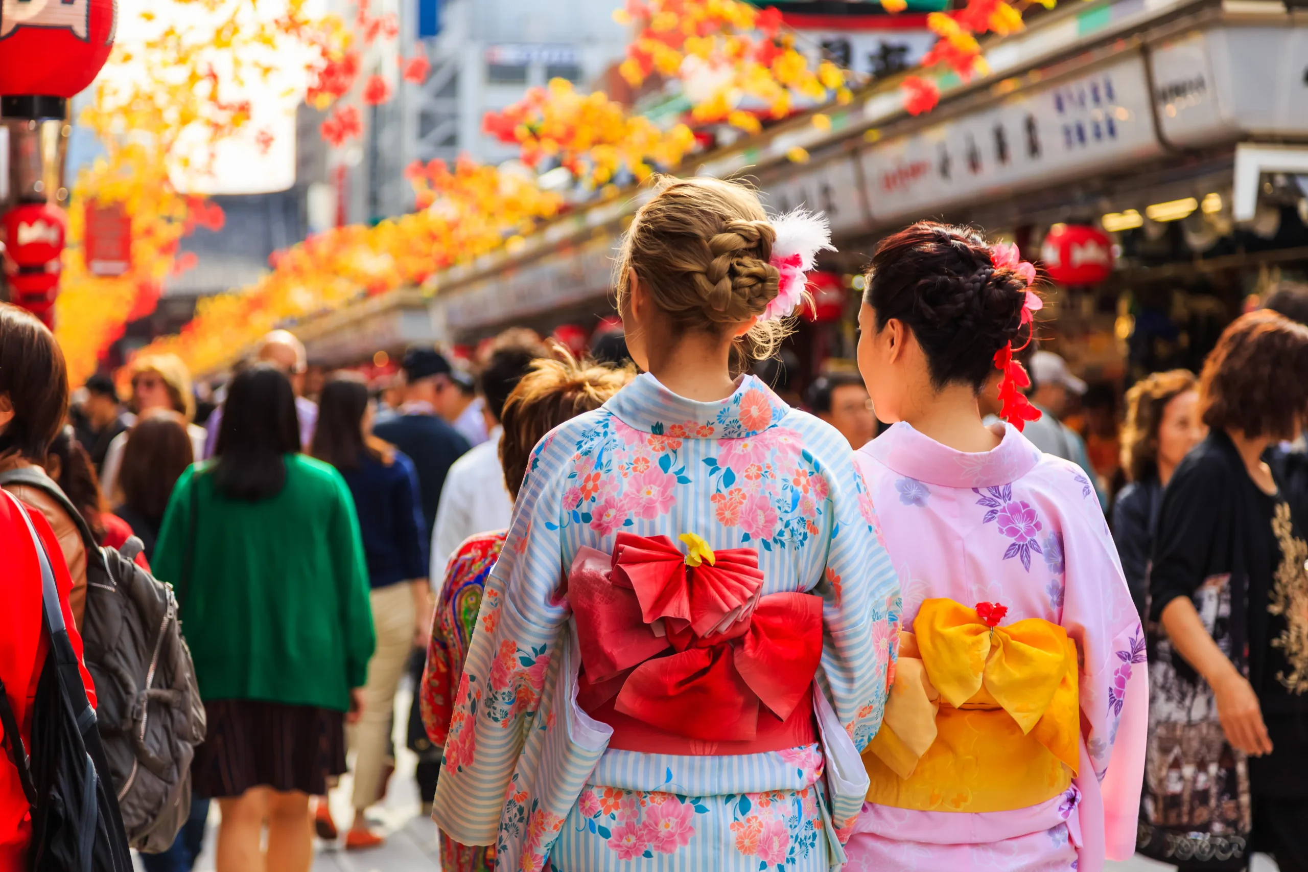 Two women dressed in colorful floral-patterned kimono, walking through a lively street adorned with festive decorations and autumn leaves.