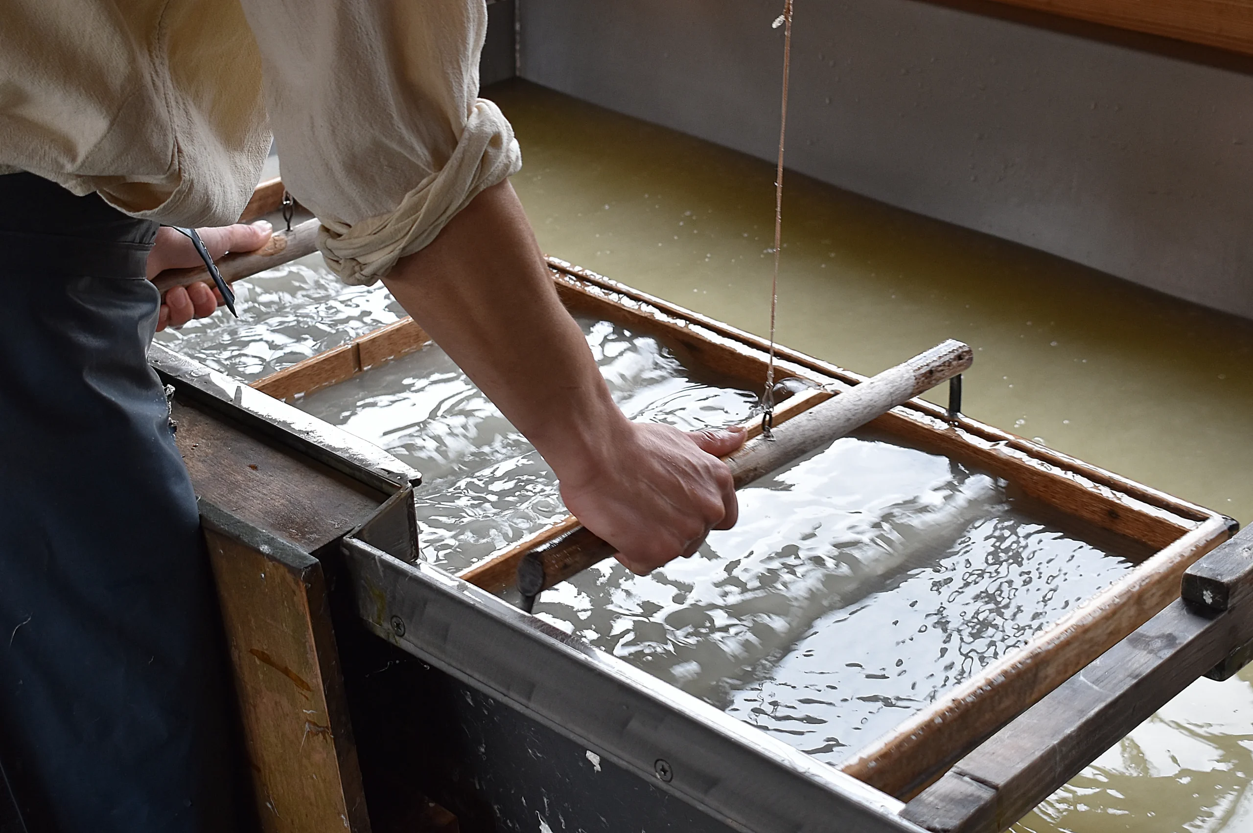 A craftsman engaged in traditional Japanese washi paper-making, carefully lifting a wooden frame from a water bath to form handmade sheets.