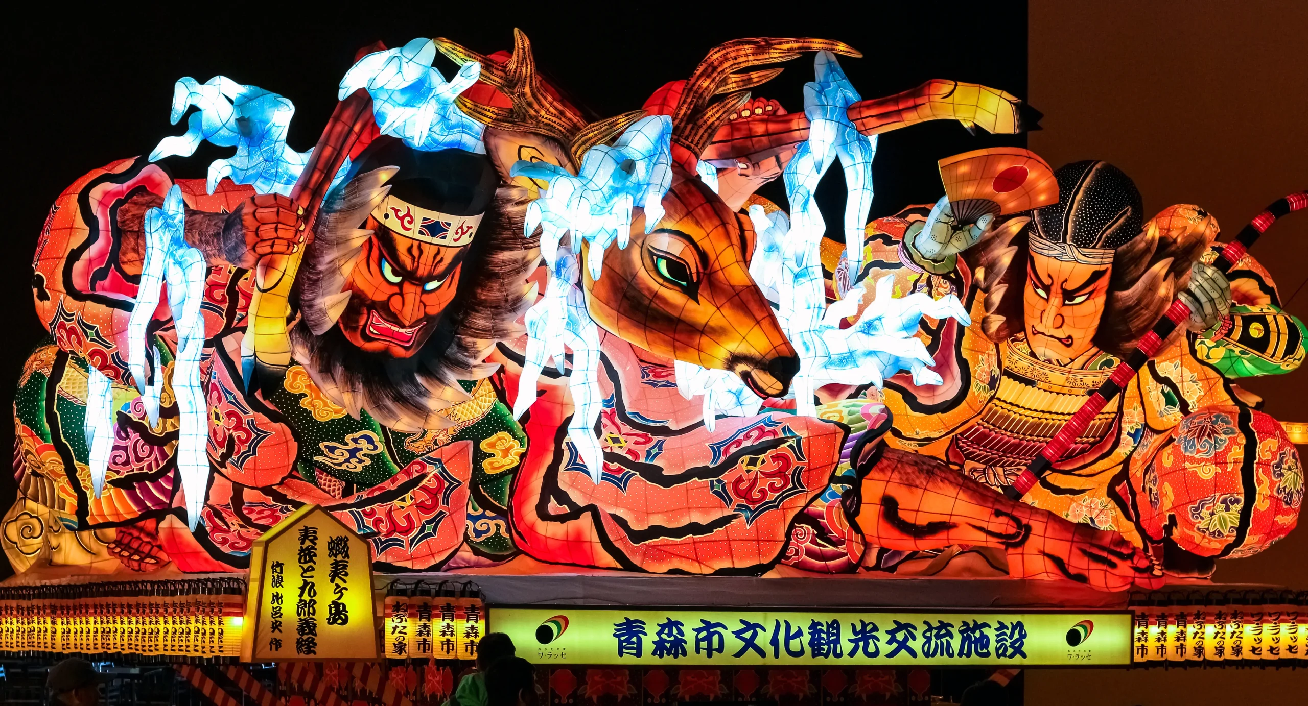 A vibrant Nebuta float from the Aomori Nebuta Festival, featuring illuminated warriors and mythical creatures with intricate details and bold colors against the night sky.