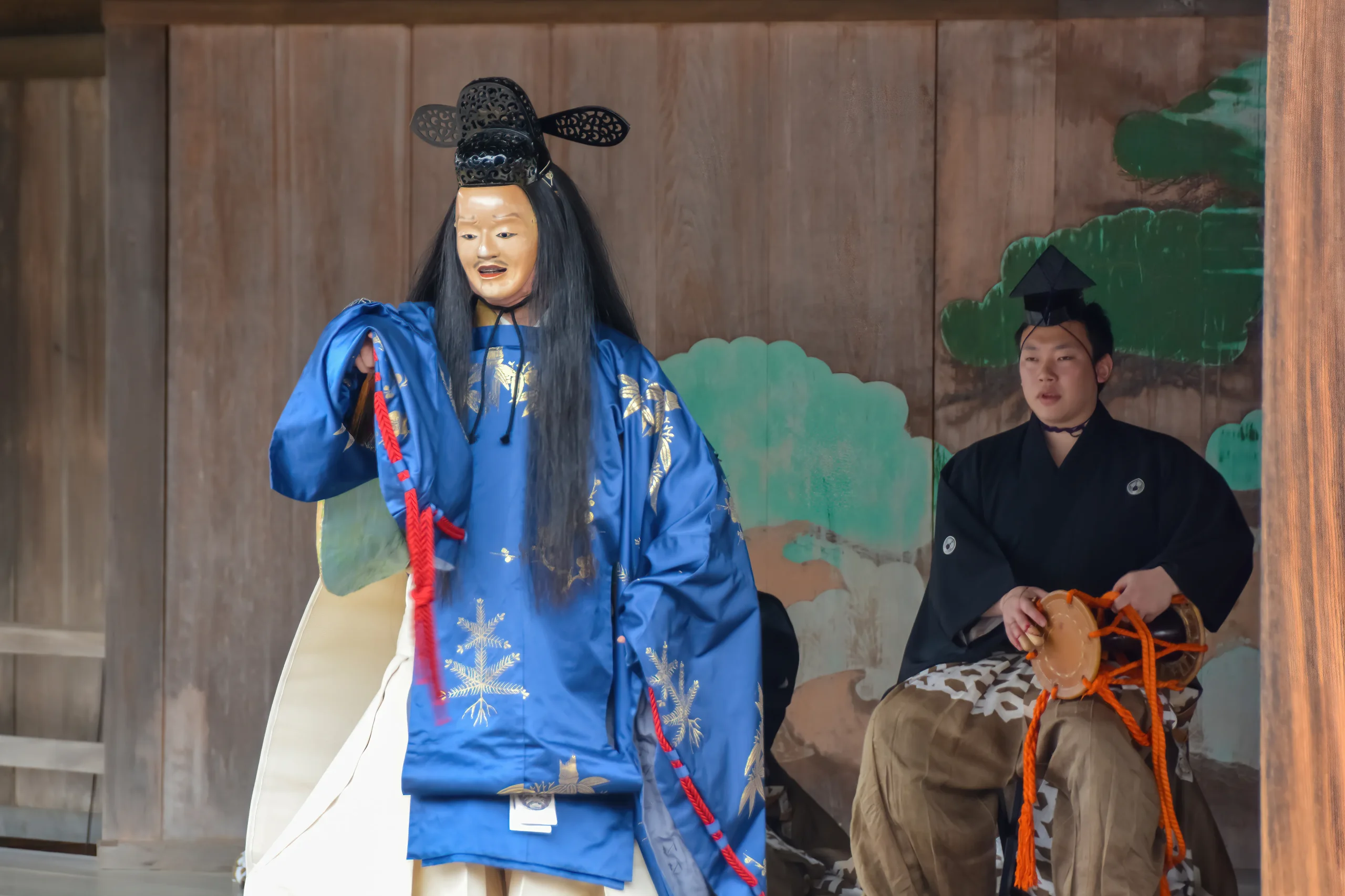 A Noh performer wearing a traditional mask and an elegant blue kimono, gracefully moving during a performance, with a musician playing in the background.