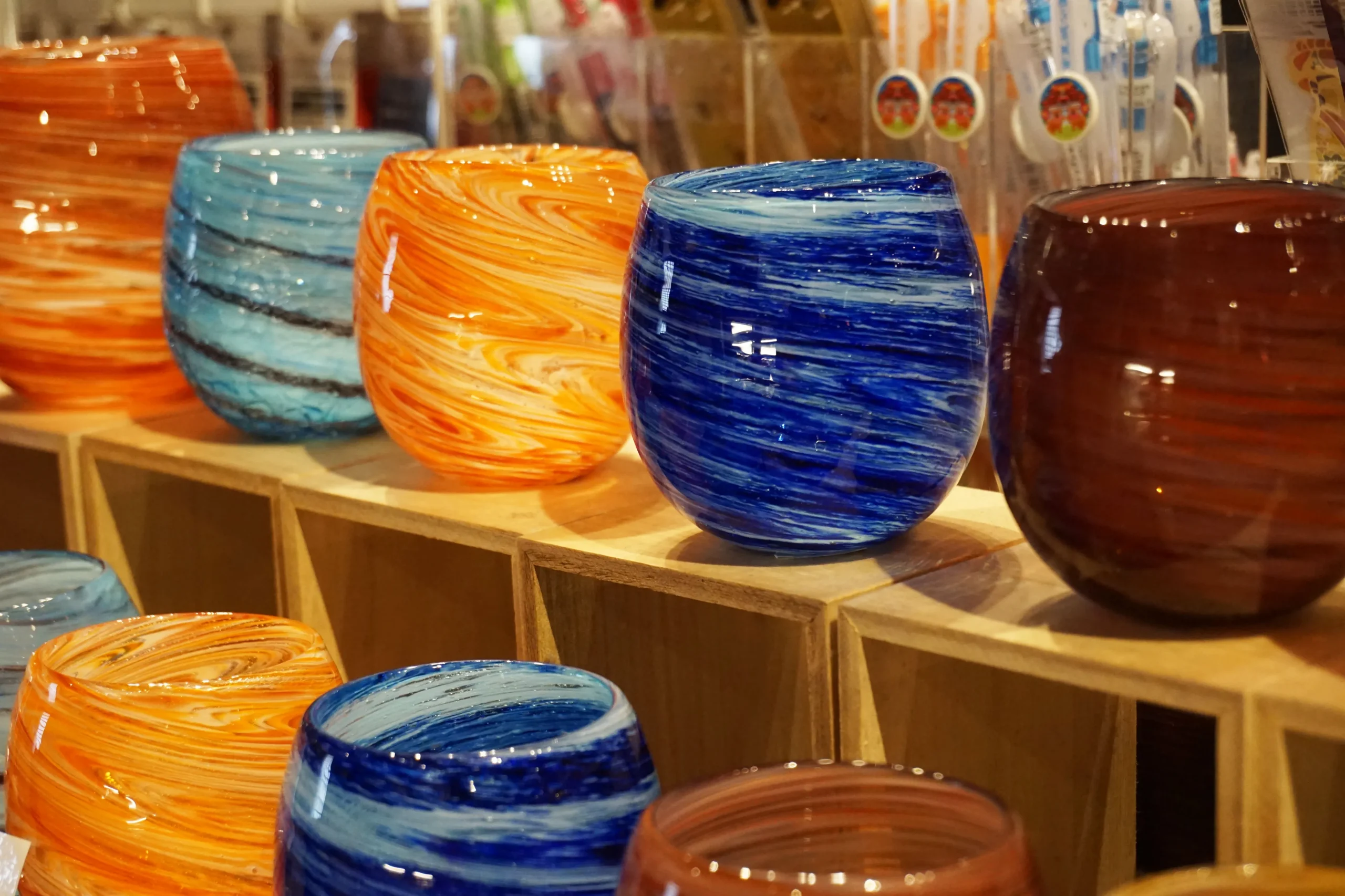 A collection of colorful, handcrafted Ryukyu glass cups on display, showcasing swirling patterns in shades of blue, orange, and red.