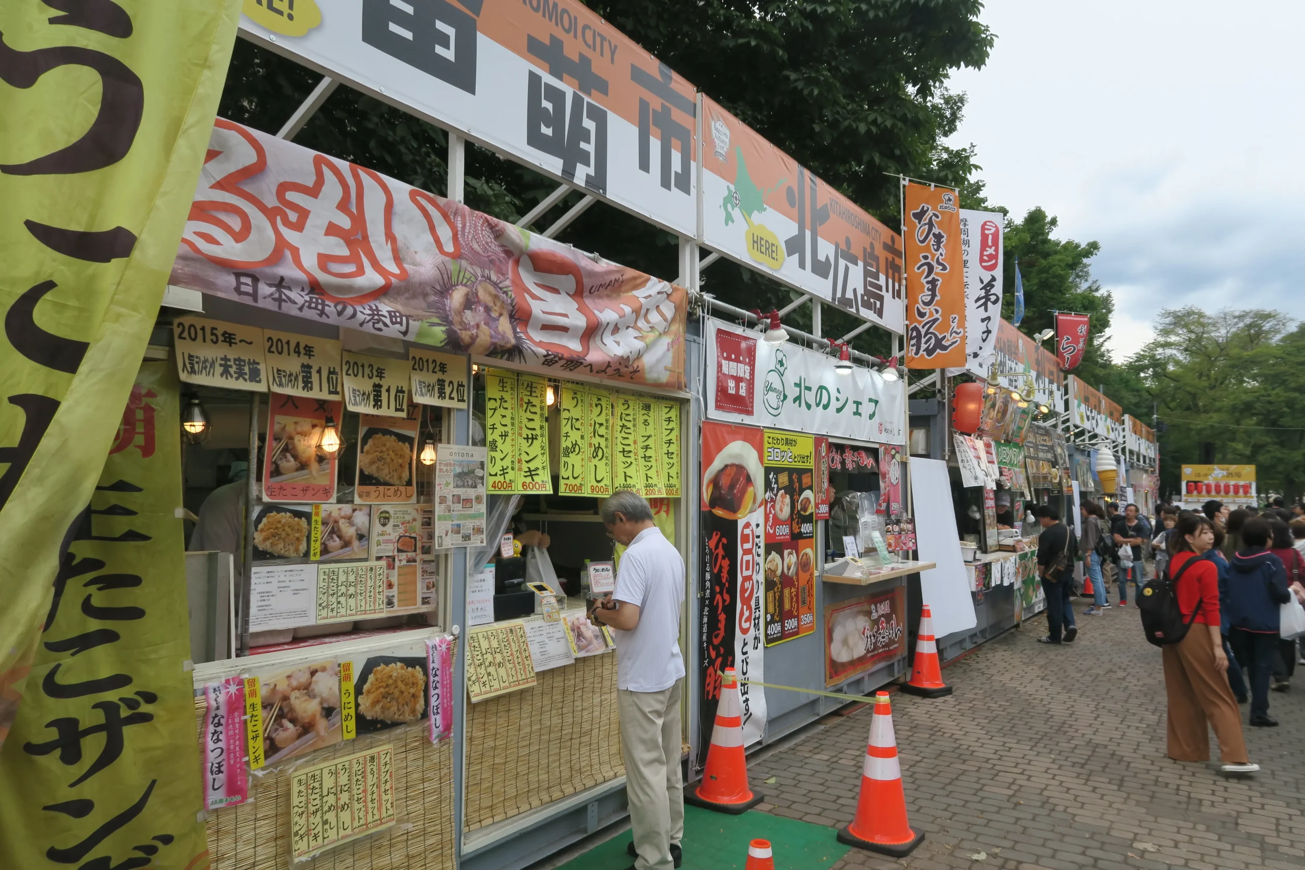 Stalls at the Sapporo Autumn Fest featuring a variety of local foods and specialties, with colorful banners, menus, and people enjoying the lively festival atmosphere in Odori Park