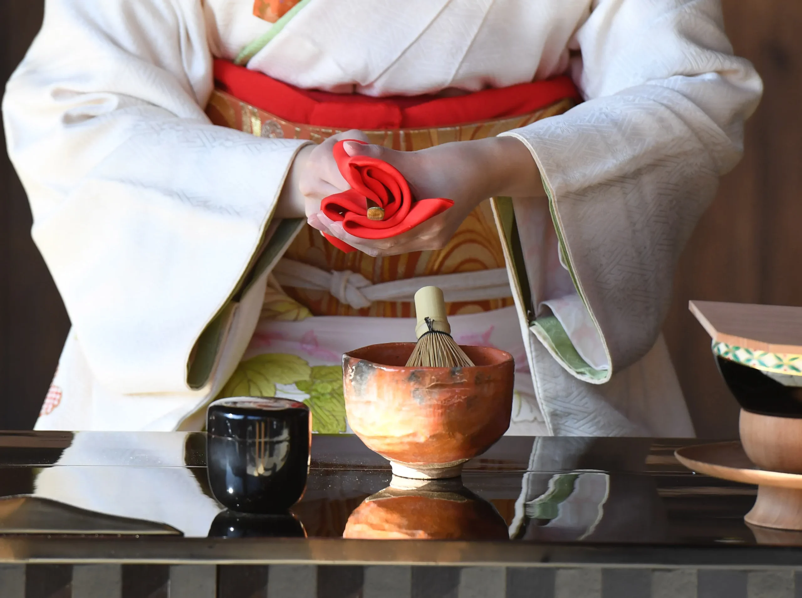 A woman dressed in a white kimono with a red obi performing a Japanese tea ceremony, delicately holding a folded red cloth, with a tea whisk and ceramic bowl on the table.