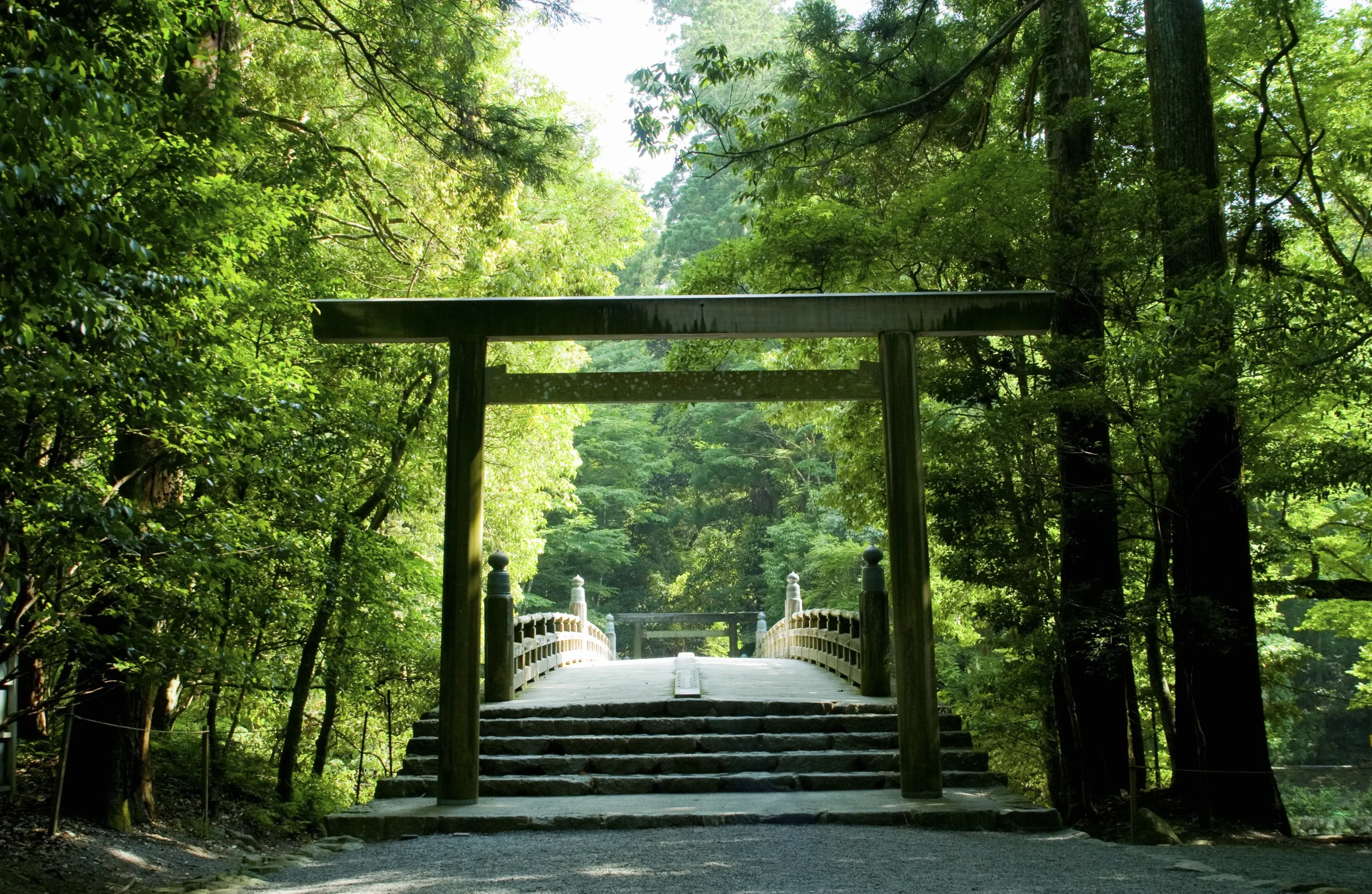 A serene wooden torii gate stands at the entrance of a sacred bridge, surrounded by lush greenery in Ise Grand Shrine, a major Shinto pilgrimage site in Japan.