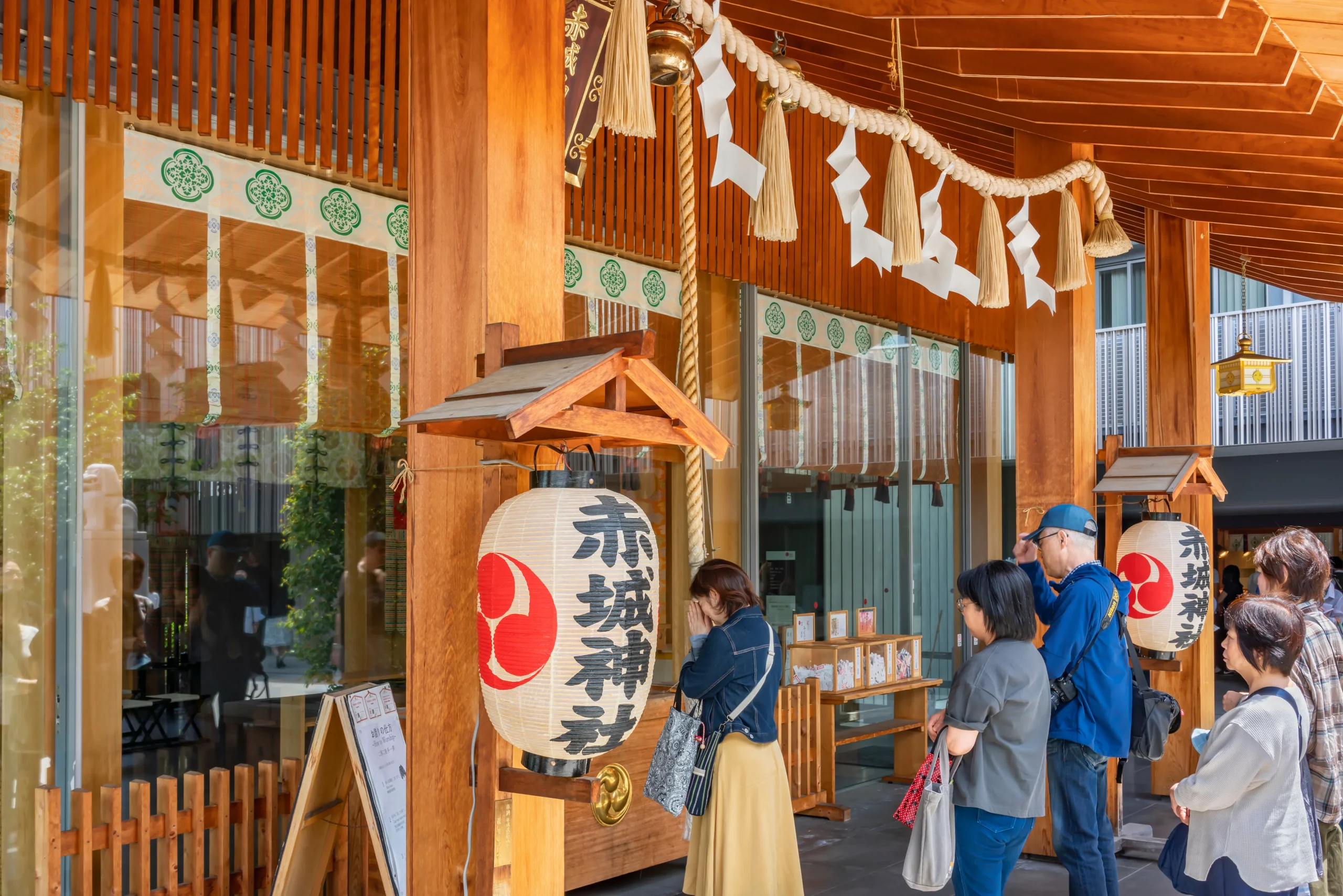 A modern wooden Shinto shrine with traditional decorations, including a shimenawa rope with shide paper strips. Visitors stand in line to pray, with a large paper lantern displaying the shrine's name.