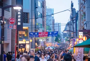 A bustling shopping street filled with festival-goers in the evening. Colorful banners, paper lanterns, and food stalls line the street, creating a lively atmosphere.