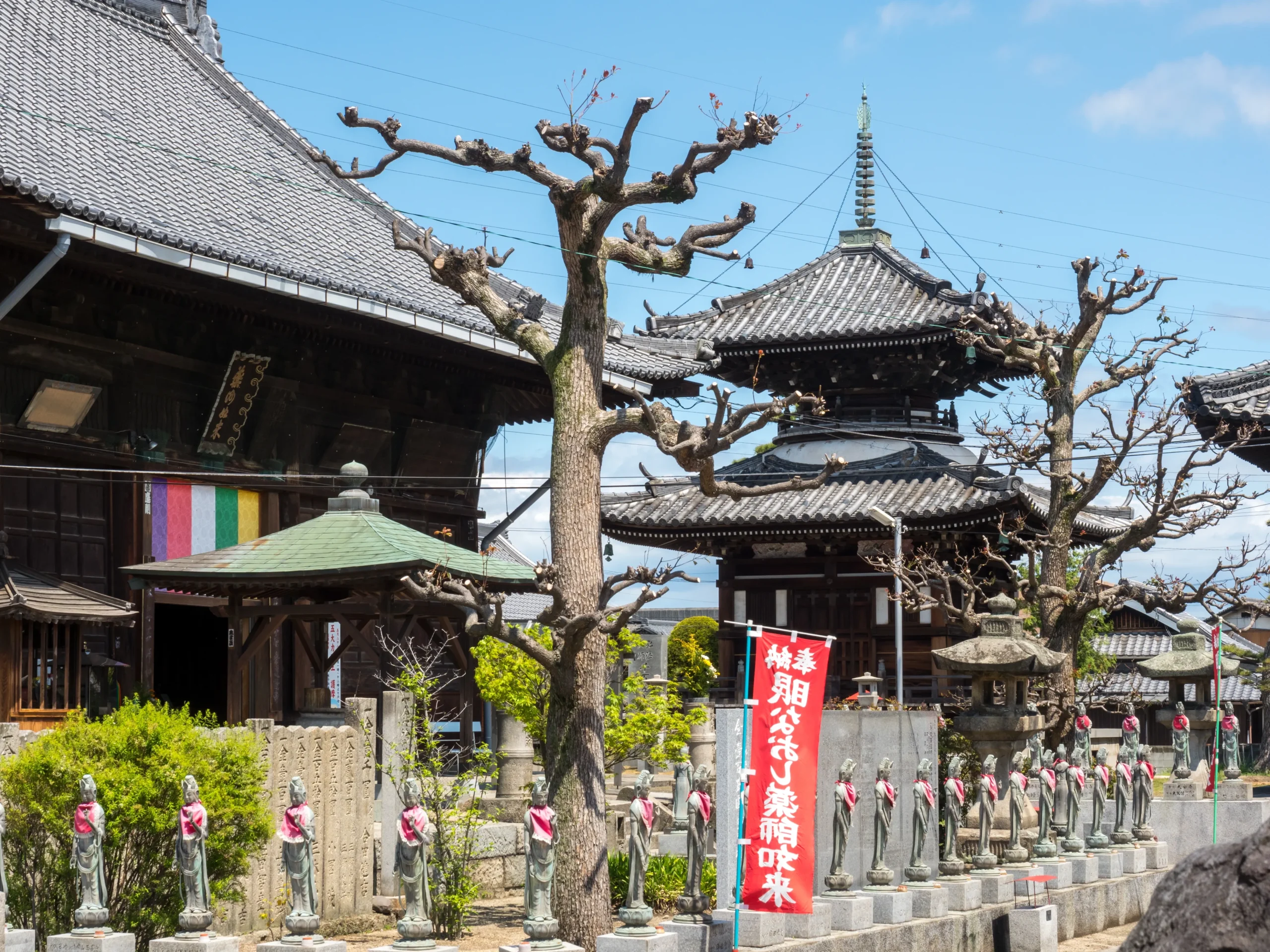 Doryuji Temple, a historic Buddhist temple featuring a traditional pagoda, wooden structures, and rows of stone Jizo statues adorned with red bibs. The temple grounds are decorated with colorful banners, and the surrounding trees have been carefully pruned, adding to the serene atmosphere.