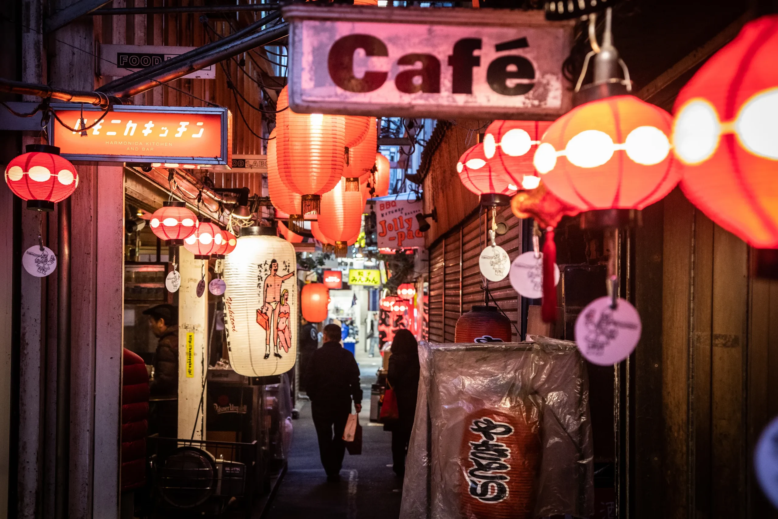 A narrow, atmospheric alley lined with izakayas and small eateries, illuminated by red paper lanterns. A mix of Japanese and English signage creates an old-school yet vibrant nightlife setting.