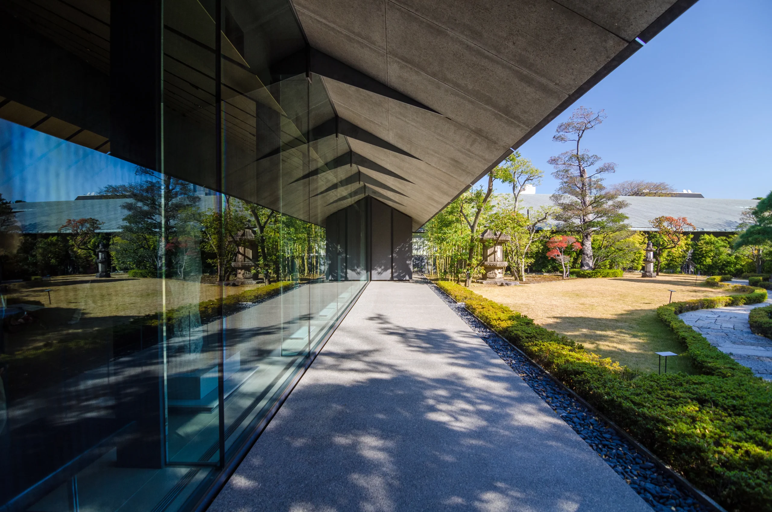 A modern architectural walkway at the Nezu Museum, featuring a minimalist concrete roof and glass walls reflecting the lush Japanese garden outside.