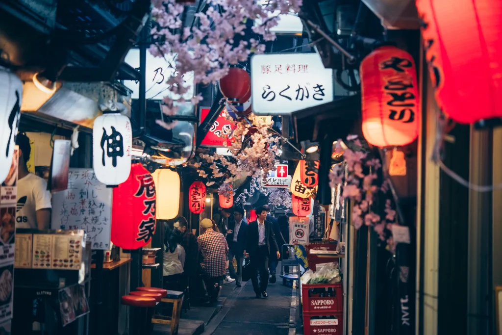 A narrow alley in Omoide Yokocho, Tokyo, lined with izakayas illuminated by red and white paper lanterns, with cherry blossoms adding to the nostalgic atmosphere.