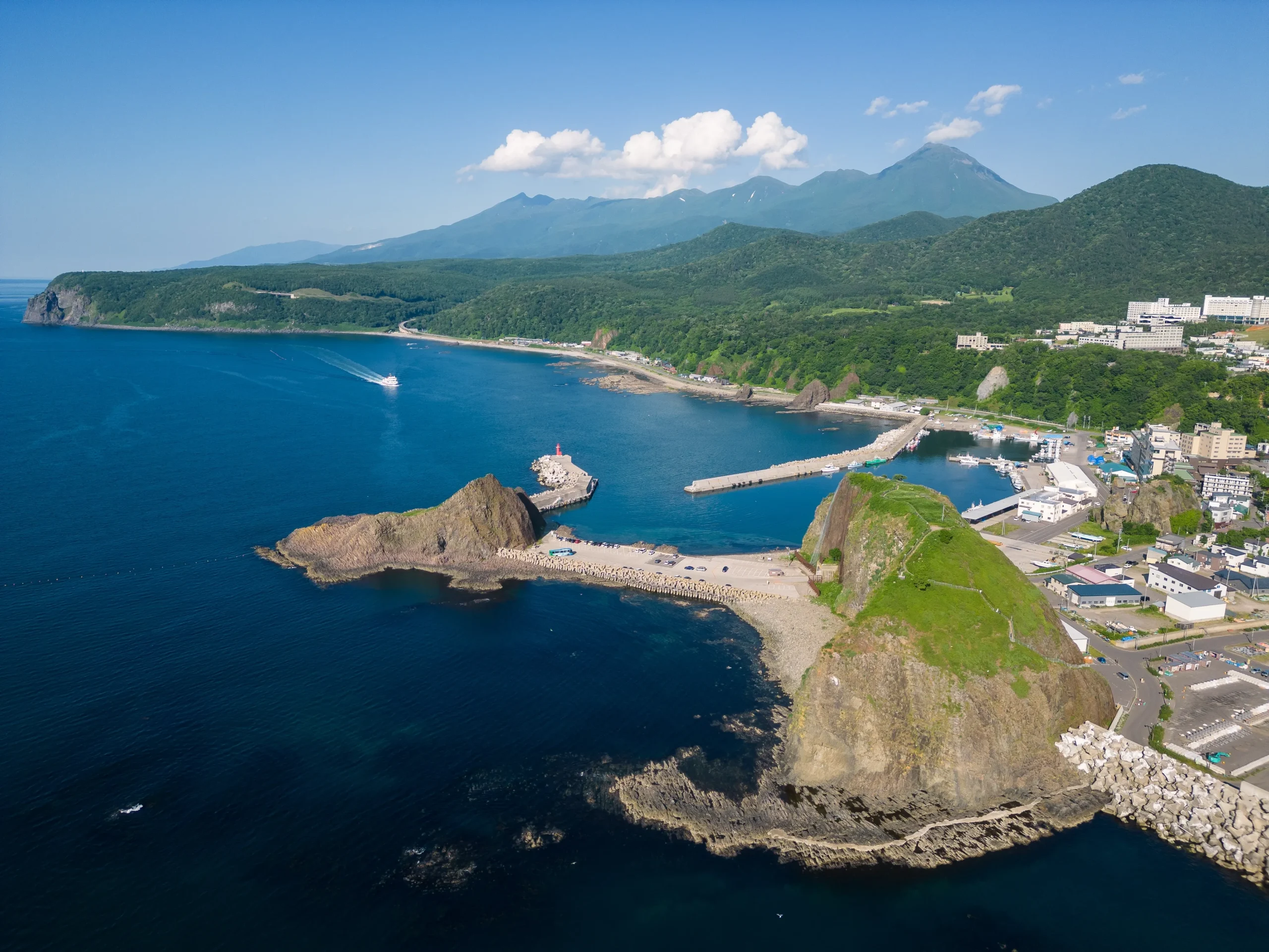 Aerial view of the Shiretoko coastline with deep blue sea, rocky cliffs, and a red lighthouse, backed by lush mountains.