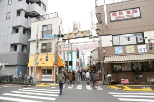 A lively entrance to Sunamachi Ginza Shopping Street, with people walking and cycling past local shops under a colorful sign with lanterns.