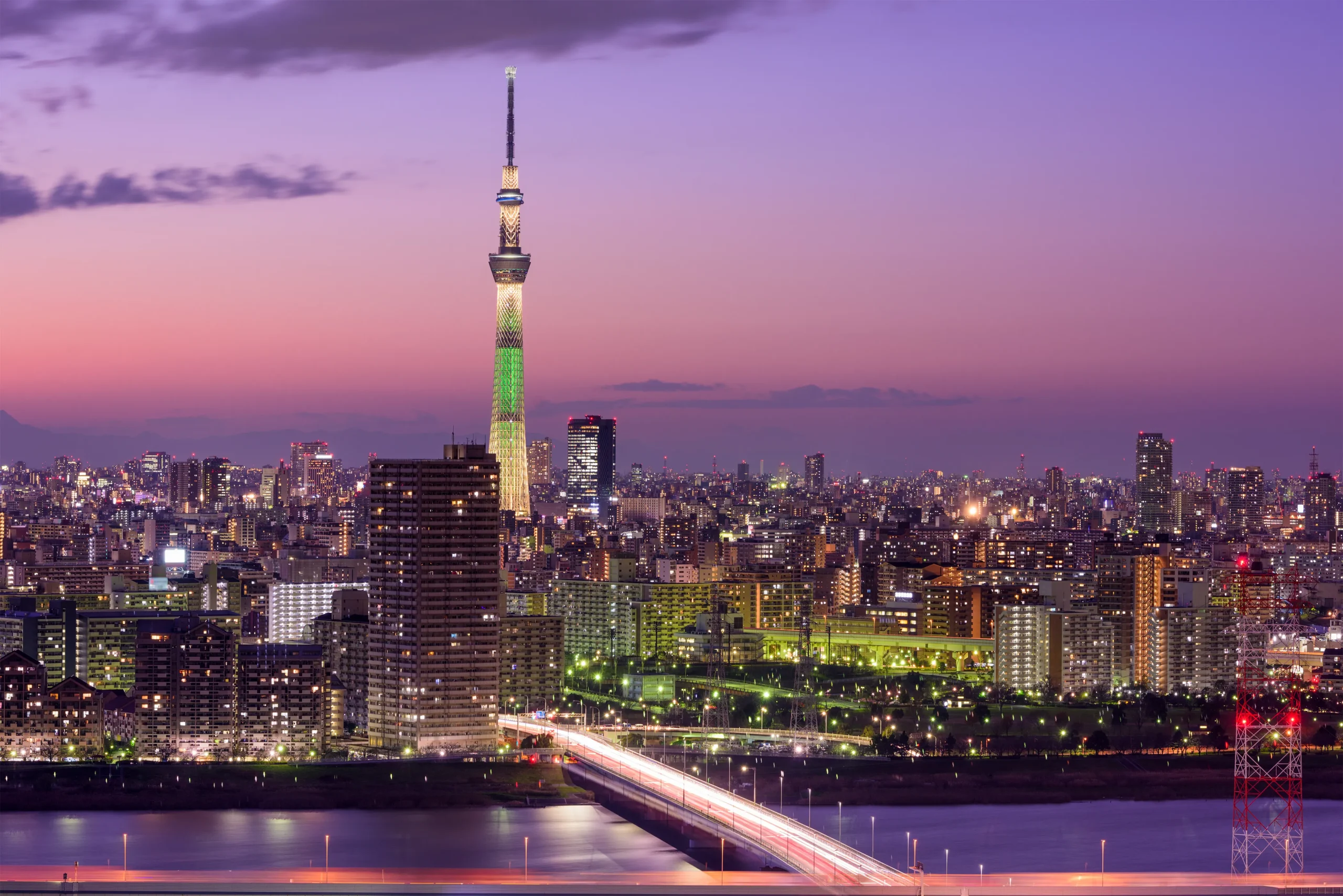 A panoramic evening view of Tokyo Skytree, brightly illuminated against a purple sky, with city lights and a river in the foreground.