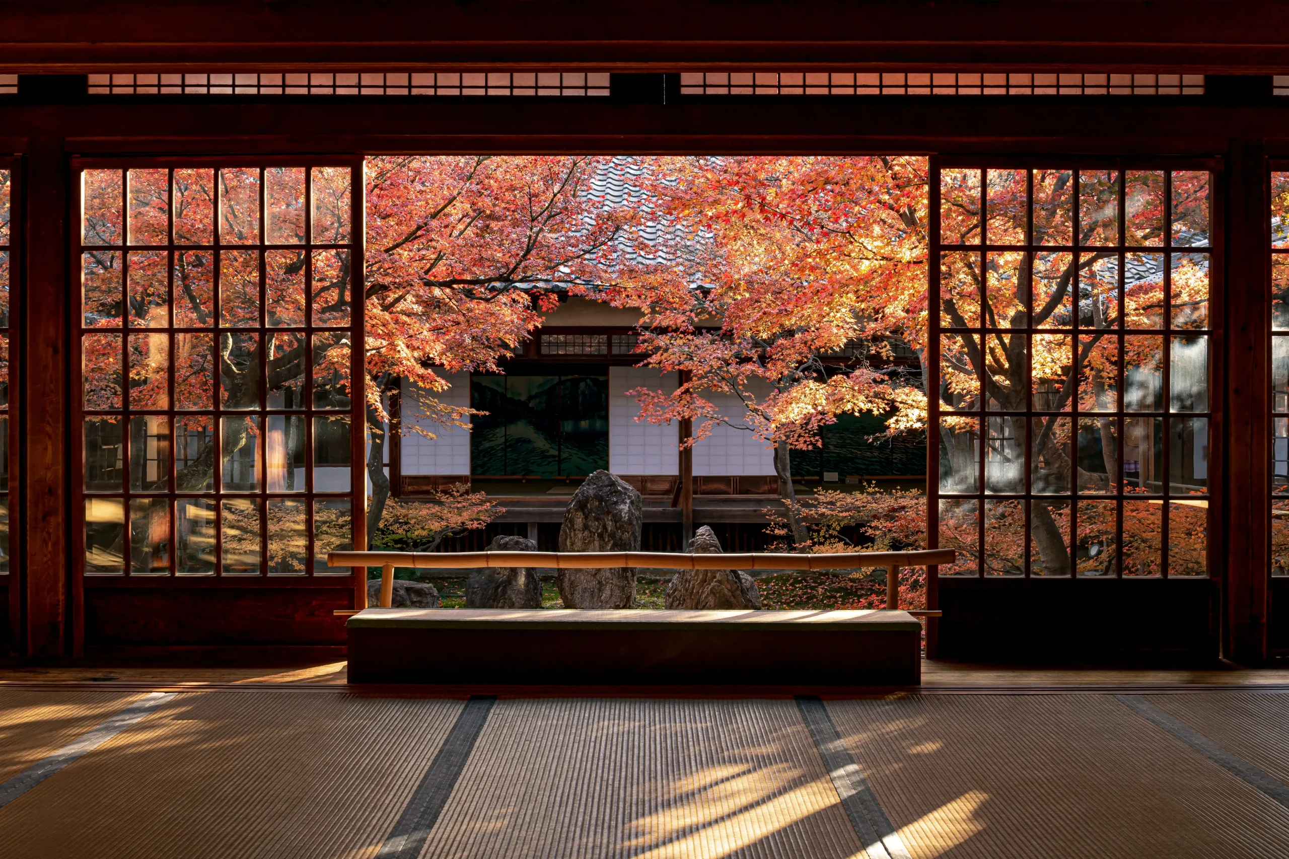 A traditional Japanese room with tatami flooring and wooden-framed glass windows, offering a breathtaking view of a courtyard garden adorned with vibrant autumn foliage.