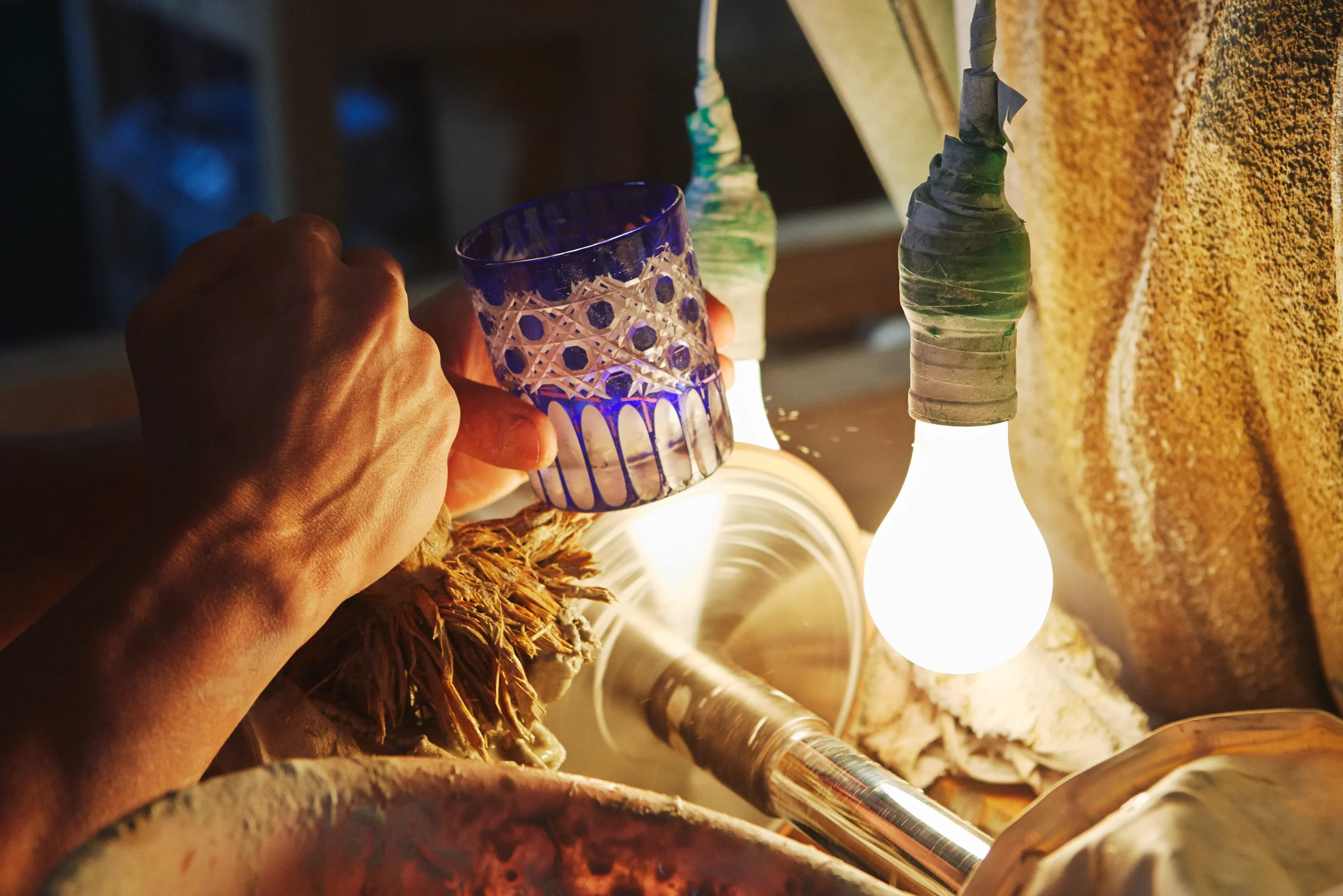 A craftsman meticulously engraving a blue Edo Kiriko glass under warm lighting, using a grinding wheel to create intricate patterns.