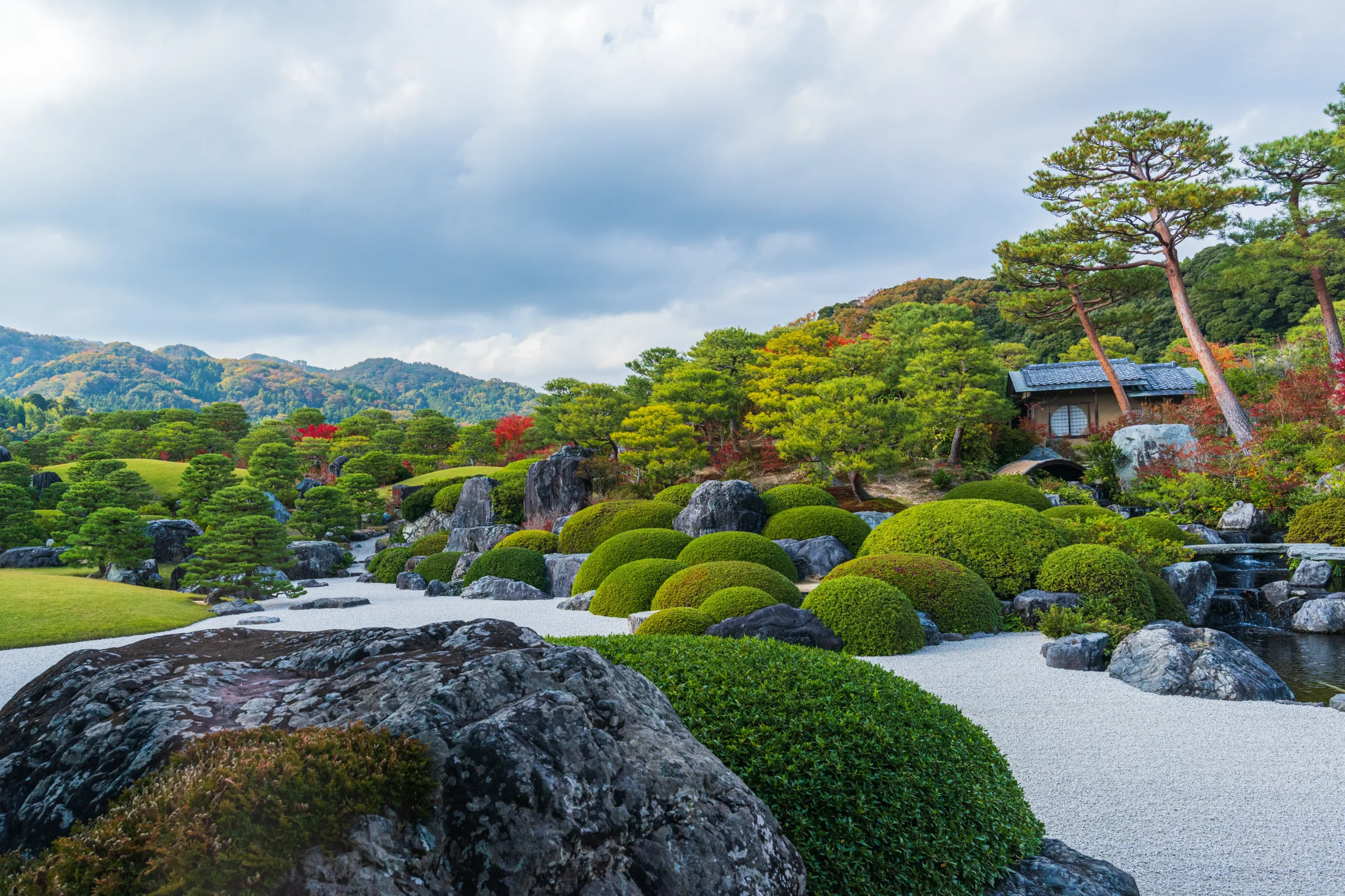 A beautifully landscaped Japanese garden featuring manicured shrubs, large rocks, and a traditional wooden tea house, with rolling mountains in the background.