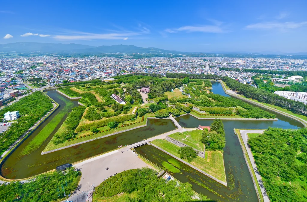 Aerial shot of Goryokaku Fort's star-shaped layout, lush with green summer vegetation, set against the city of Hakodate.