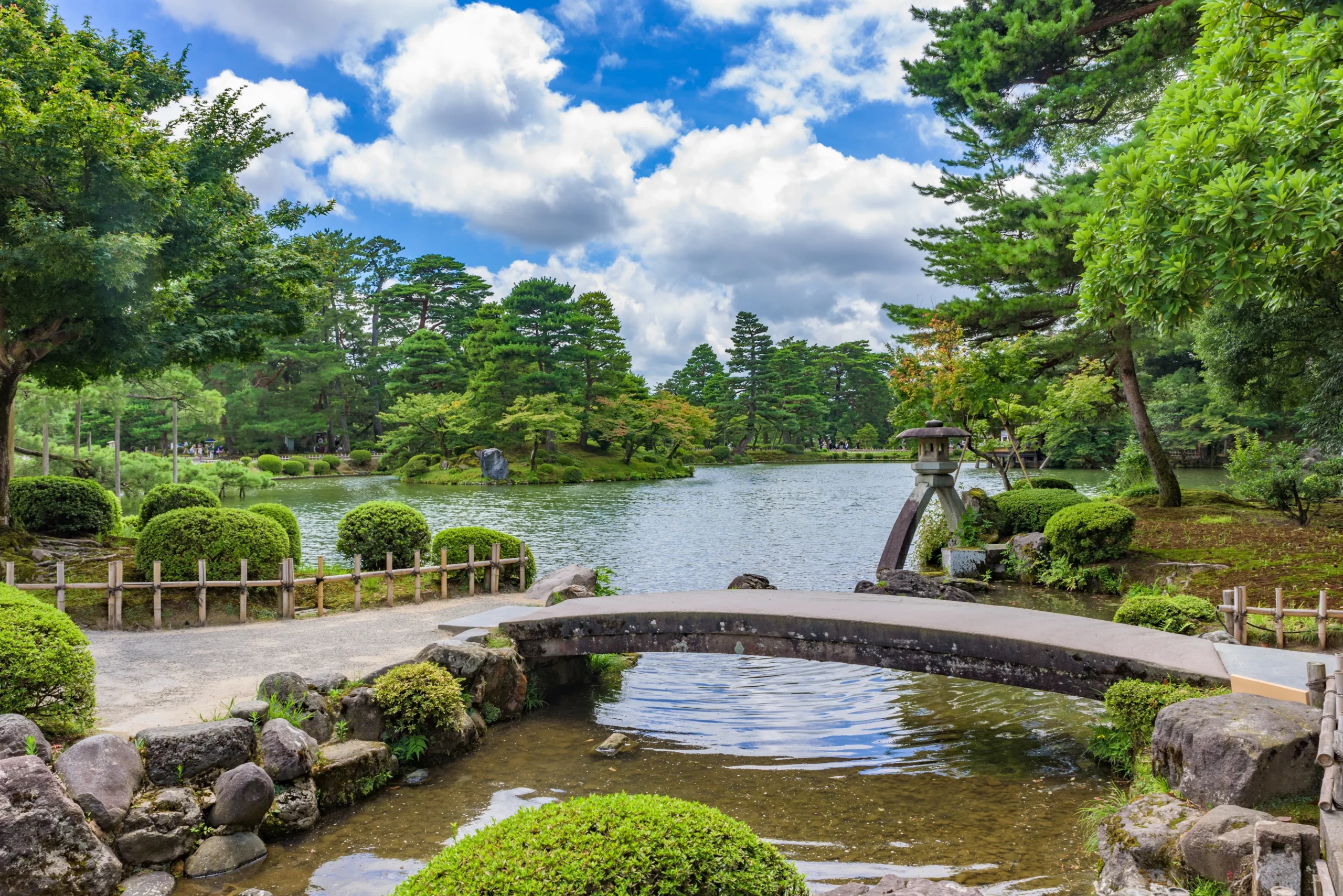 A scenic Japanese garden with a tranquil pond, a curved stone bridge, and a traditional stone lantern, surrounded by lush greenery and towering pine trees.