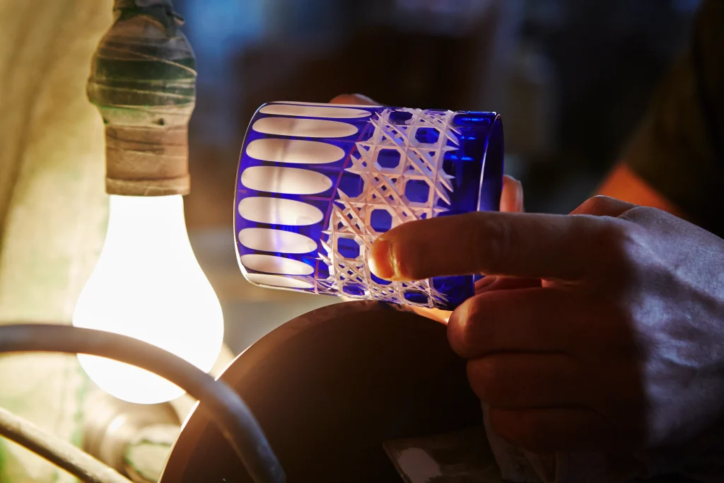 A craftsman carefully refining the edges of an Edo Kiriko glass, holding it against a rotating grinding wheel under the glow of a work lamp.