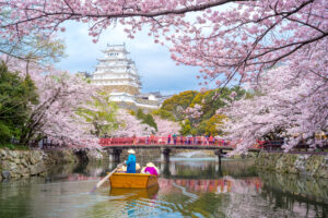 A traditional boat ride under full-bloom cherry blossoms, with Himeji Castle in the background and a red bridge spanning the moat.