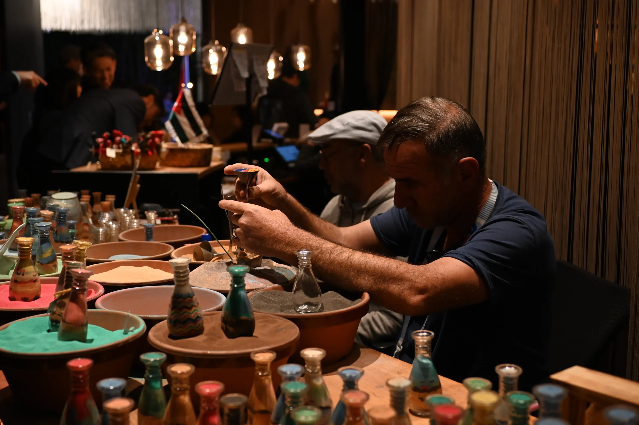 An artisan carefully creating colorful sand art in glass bottles at an indoor booth, surrounded by vibrant sands and jars.