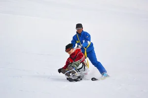 An instructor guiding a seated skier on adaptive ski equipment down a snowy slope at Fujimi Kogen Ski Resort.