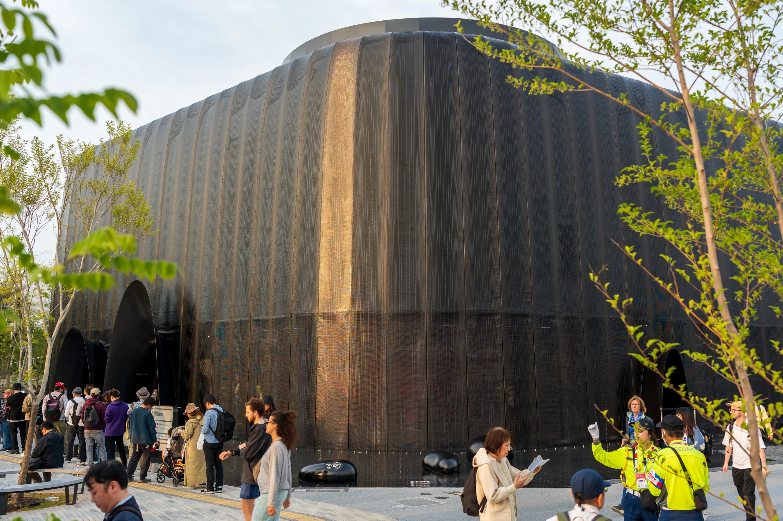 Crowds gather outside a dark, futuristic dome-like pavilion surrounded by trees at Expo 2025.