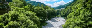 A clear riverbed lined with pebbles, surrounded by dense green forest and mountain villages in the distance.