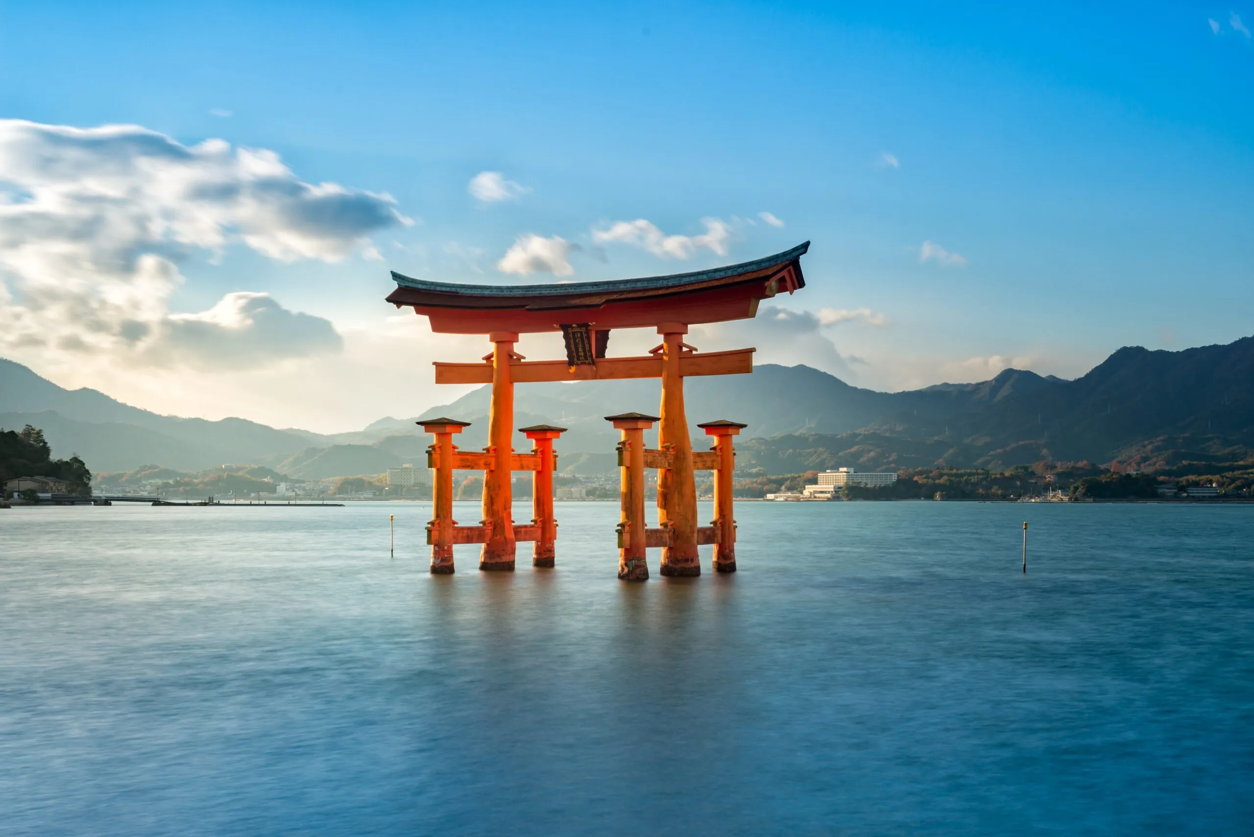 The floating red torii gate of Itsukushima Shrine standing in the sea with a backdrop of mountains and blue sky.