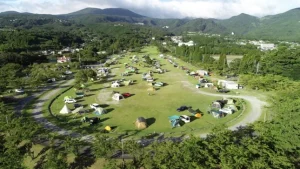 An expansive grassy campground in a mountainous area with scattered tents and vehicles under a partly cloudy sky.