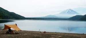 A solo lakeside campsite with a tarp tent, fishing gear, and Mount Fuji in the distance across the calm water.