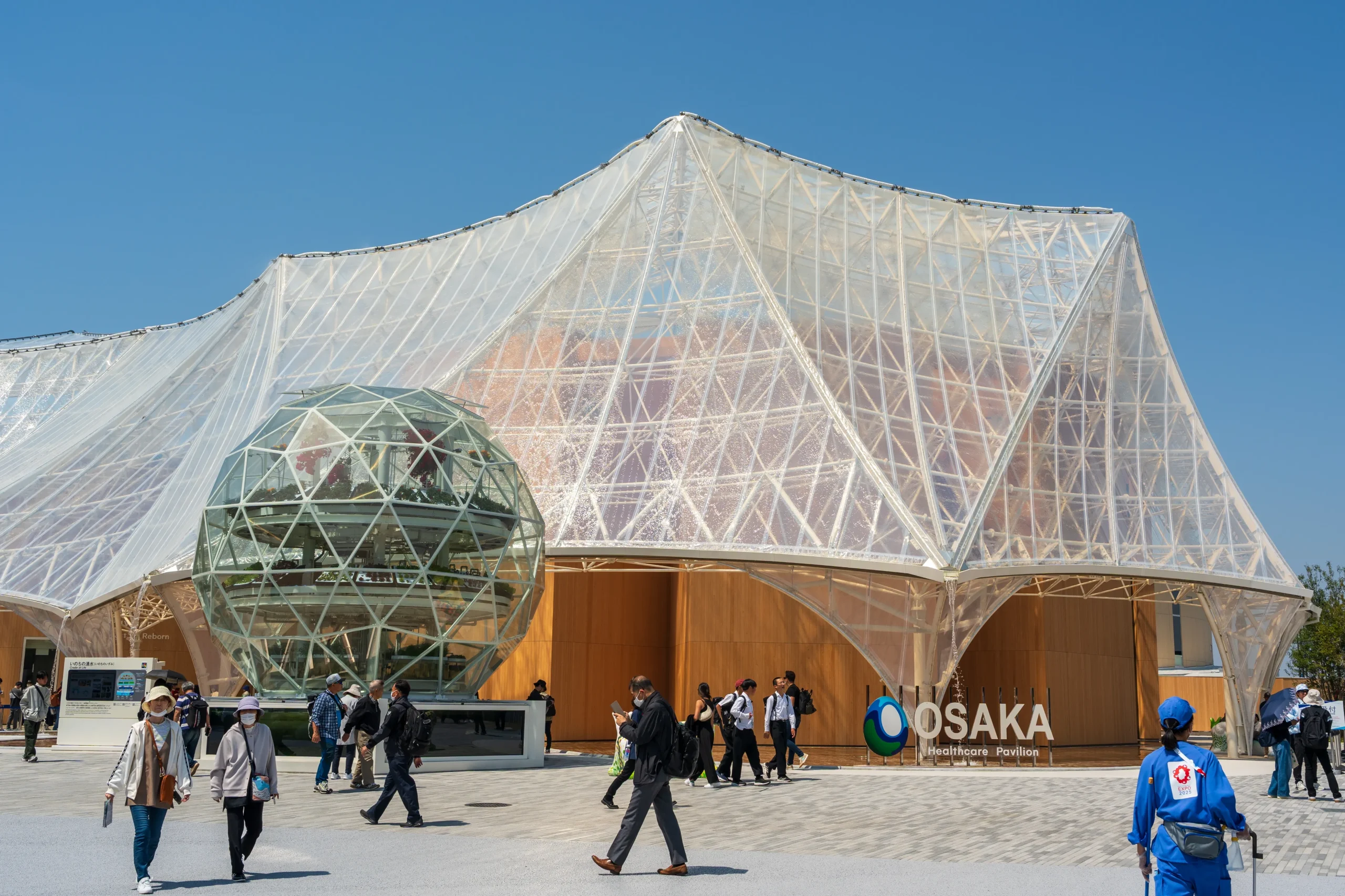 Glass dome and airy architectural structure of the Osaka Healthcare Pavilion, with visitors walking nearby.